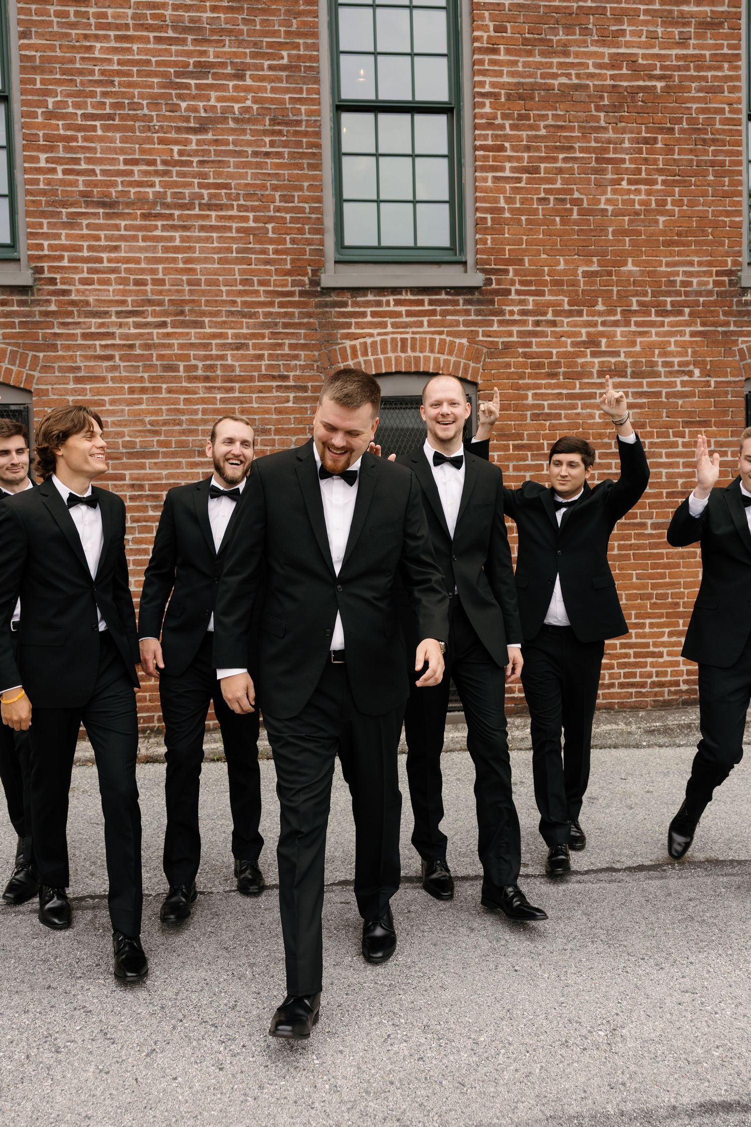 A line of groomsmen in black tuxedos lean sideways in a coordinated pose against a brick wall background.