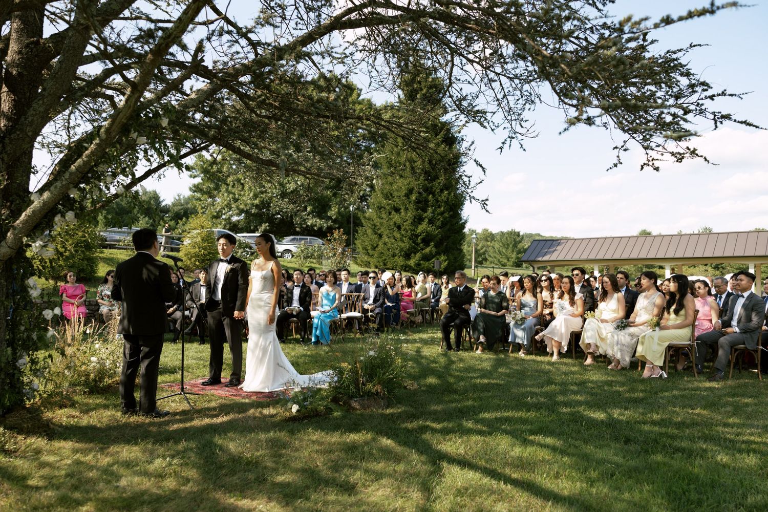 Outdoor wedding ceremony under trees with guests seated on white chairs.