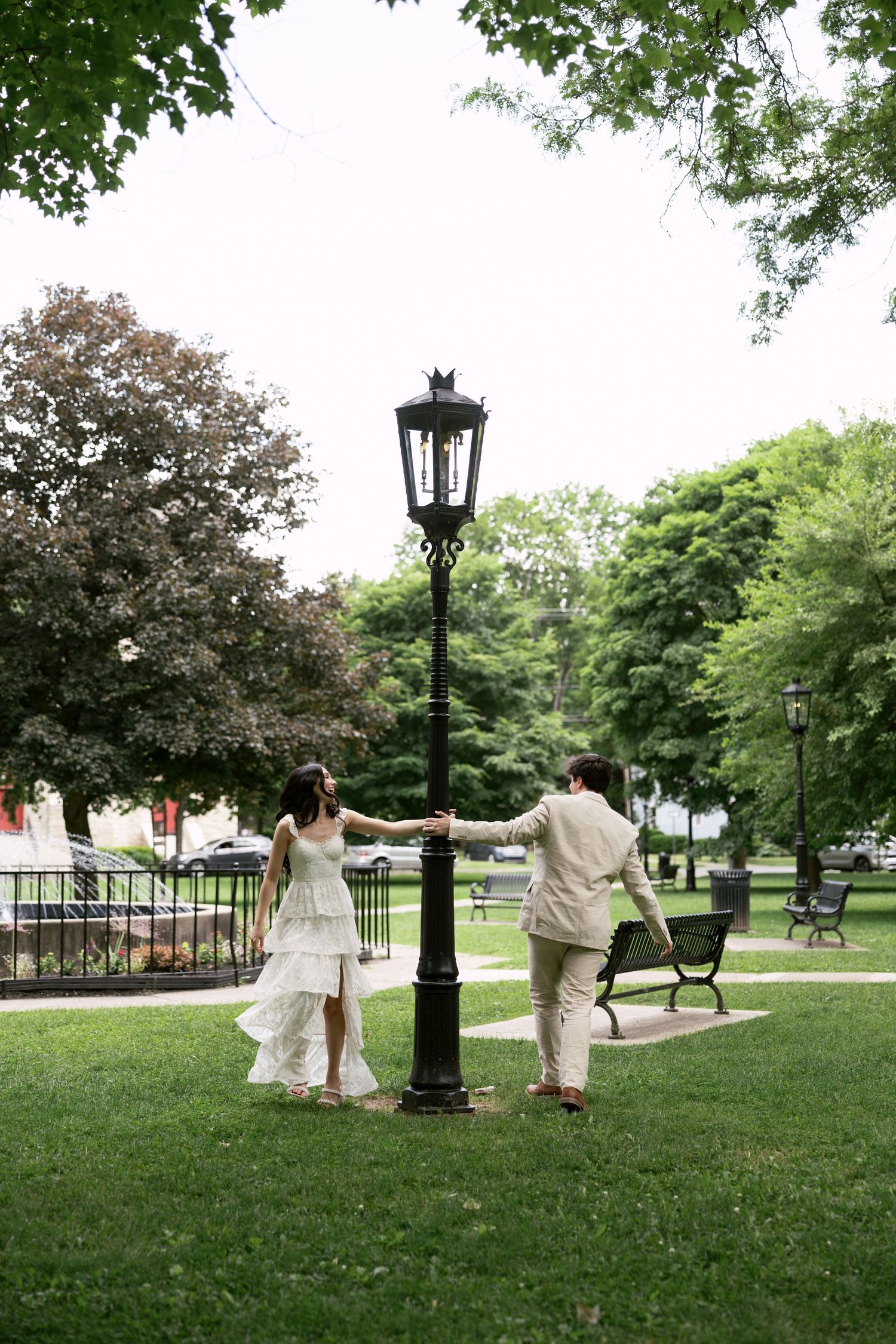 A couple in wedding attire walks along a scenic park path with trees and lamp posts.