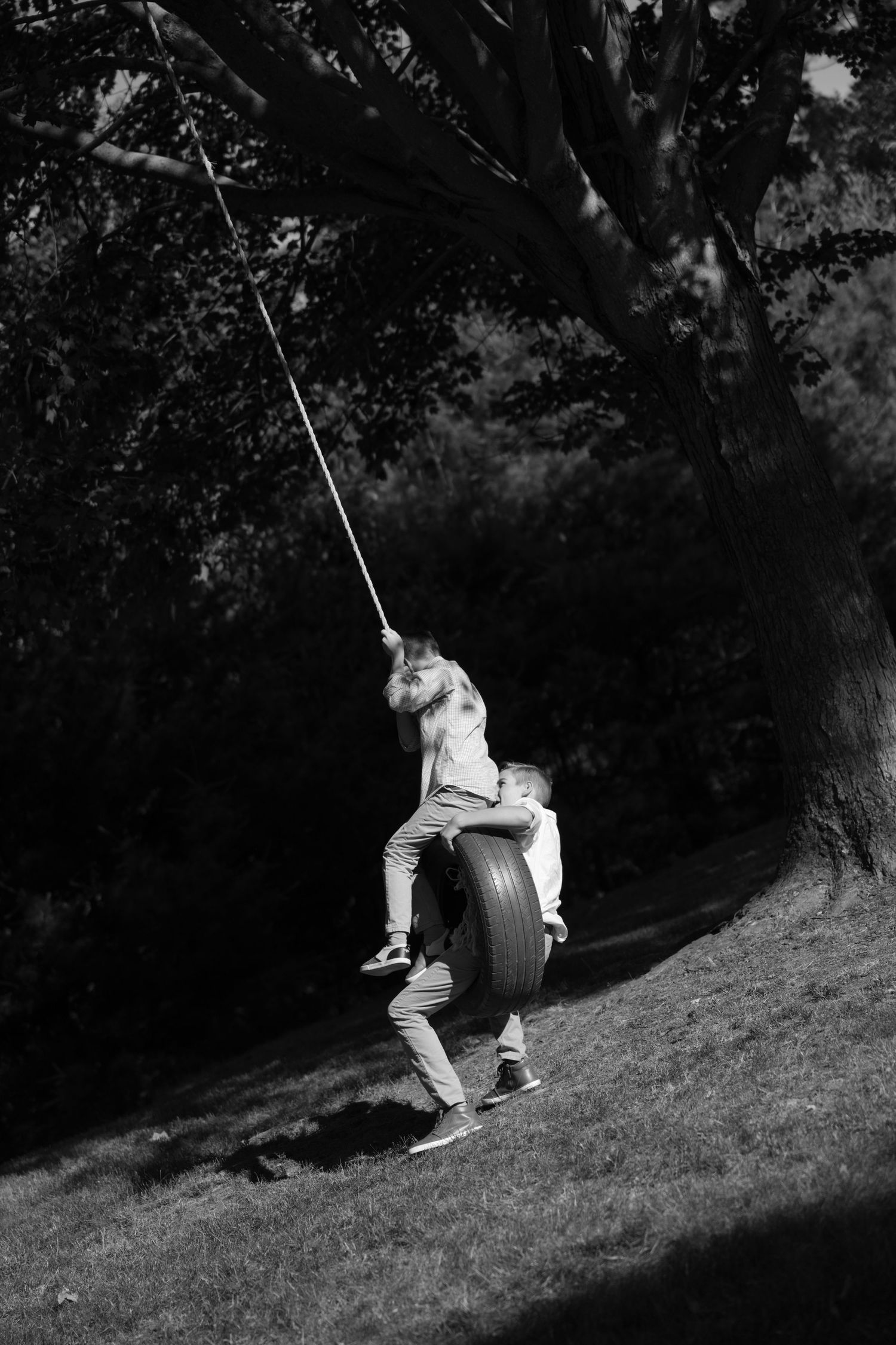 Black and white photo of someone enjoying a tire swing in a shaded outdoor setting.