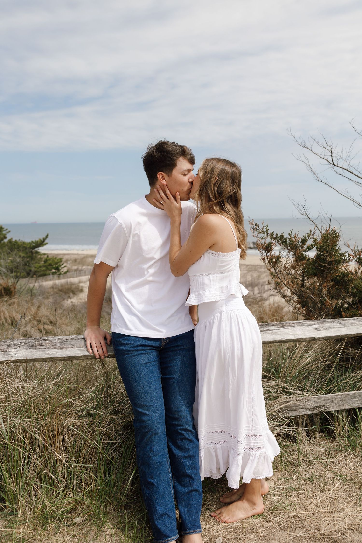 A couple in casual white outfits sharing romantic moments along a coastal boardwalk.