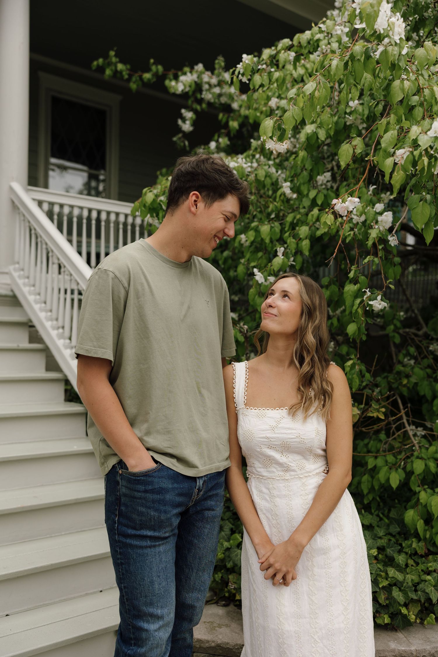 A couple sharing an intimate moment against white flowering bushes and a house exterior.