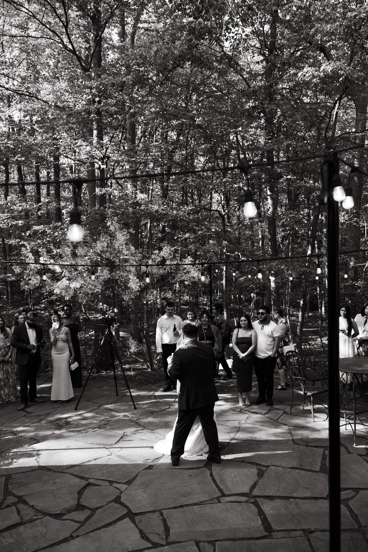 Black and white photo of a wedding ceremony taking place outdoors with trees and string lights in the background.