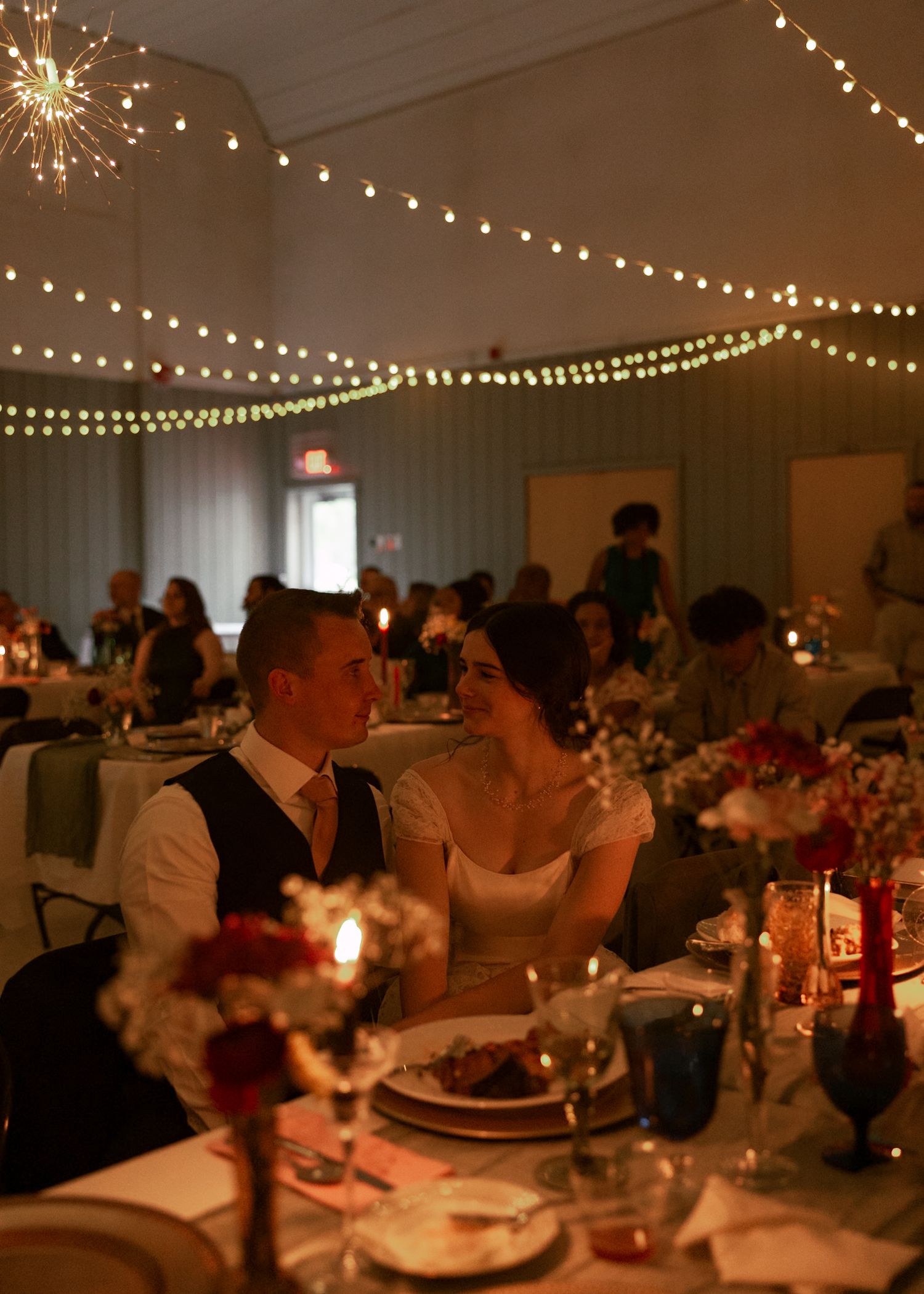 Wedding reception venue with string lights and tables set for dinner in a dimly lit room.