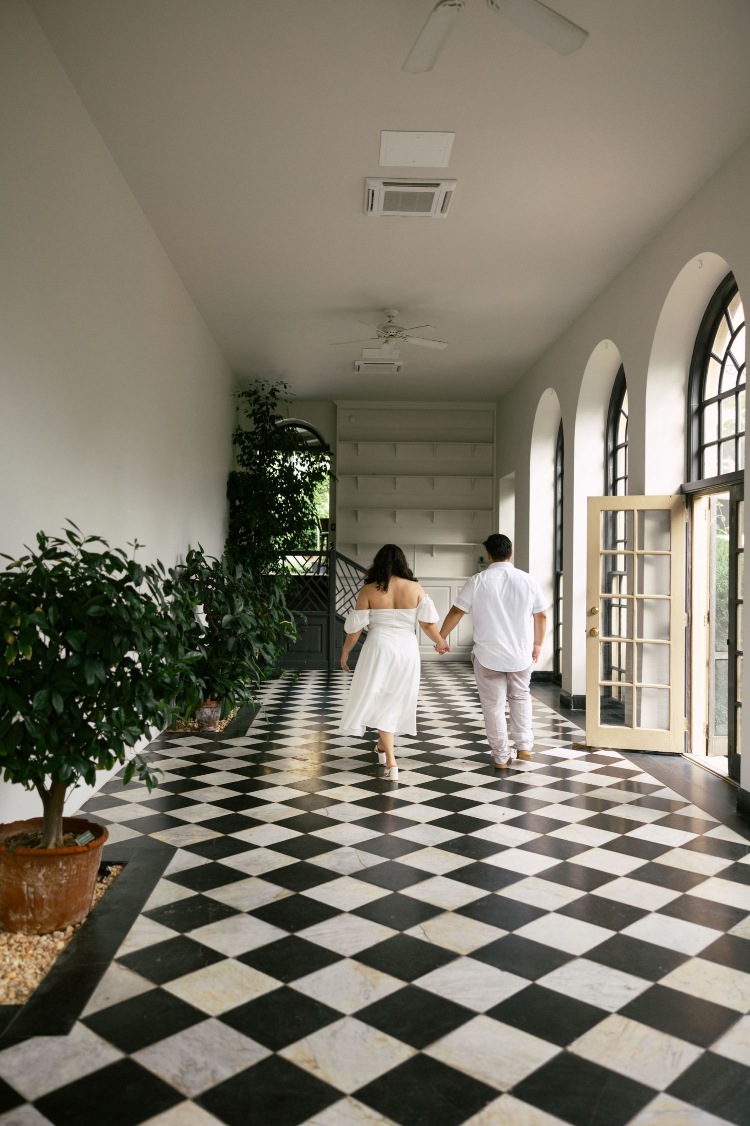 A long hallway with black and white checkered floor tiles features arched windows and potted plants along the wall.