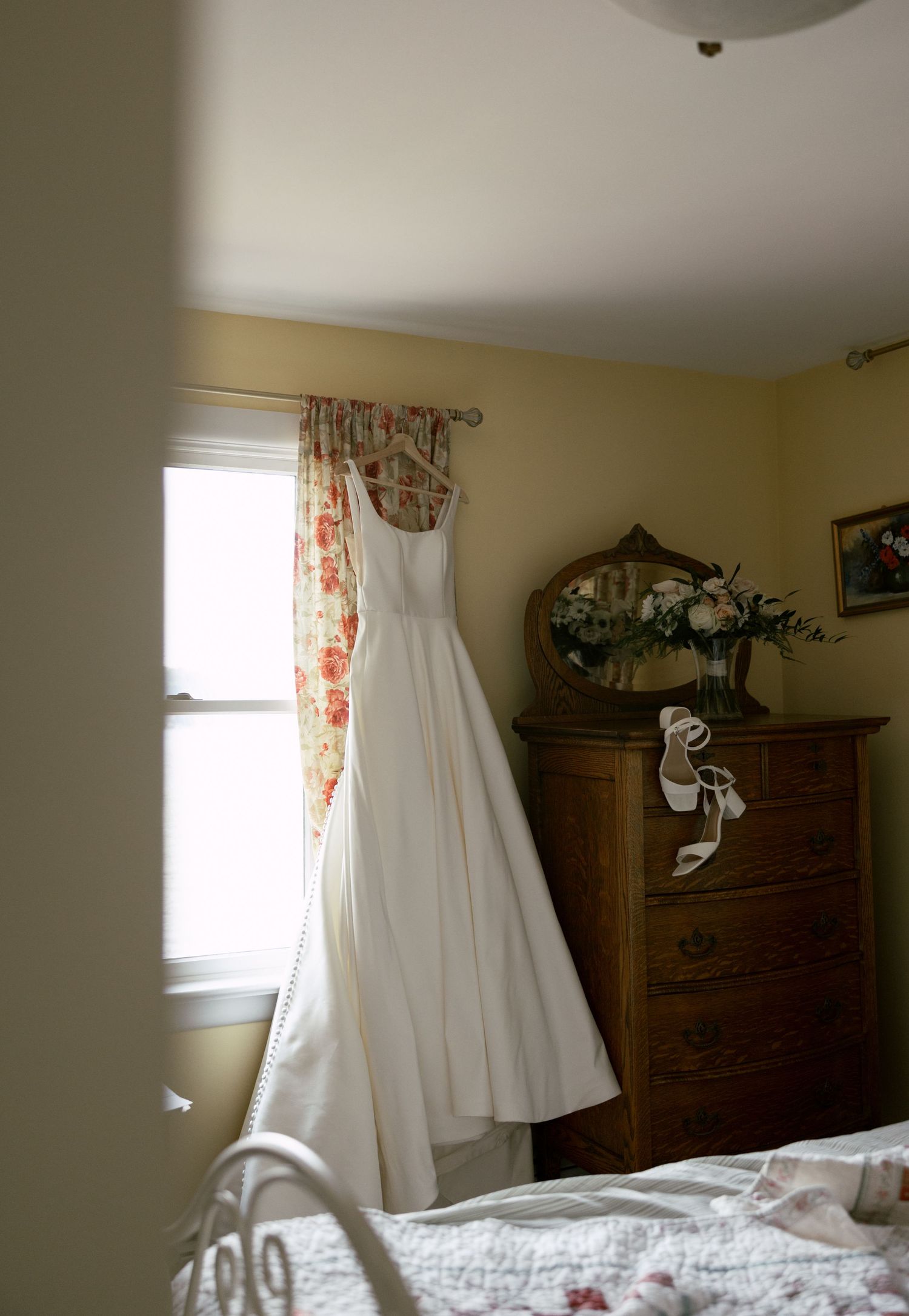 A white wedding dress hangs elegantly in a sunlit bedroom with floral curtains.