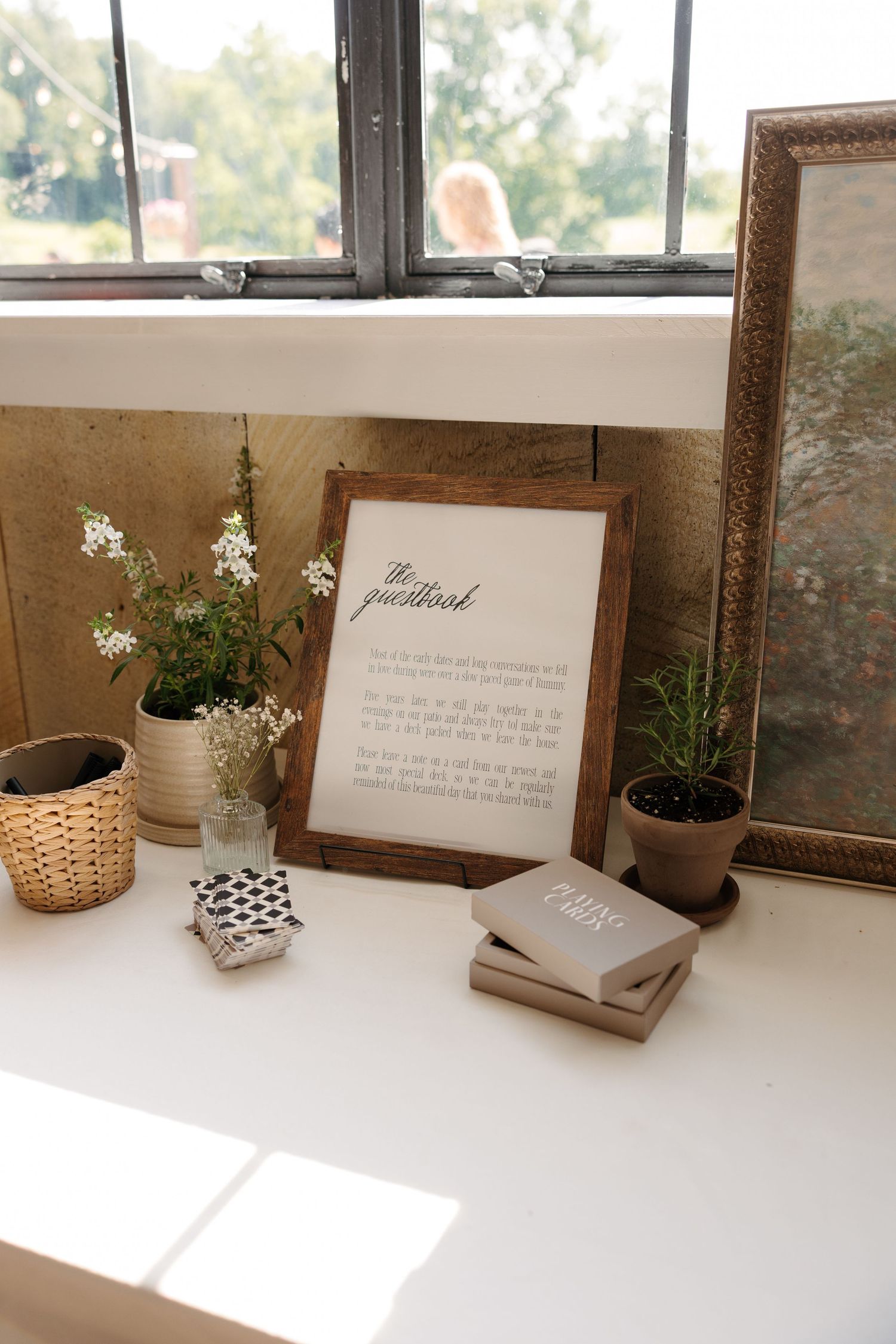Rustic wedding guestbook display with framed sign, wildflowers, potted plants, and books on a white windowsill.
