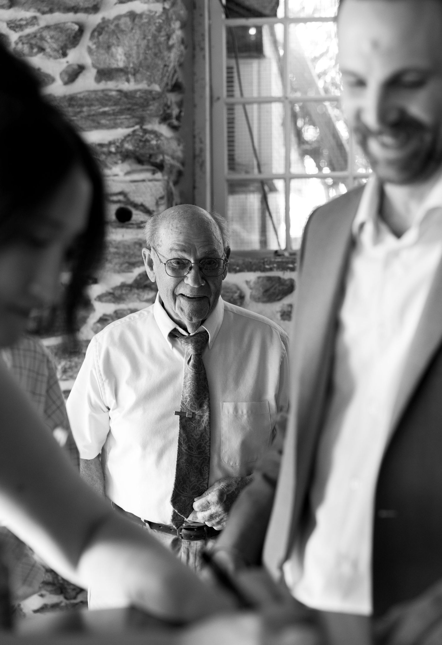 Elderly man in glasses and tie smiling warmly at a social gathering in black and white photo.