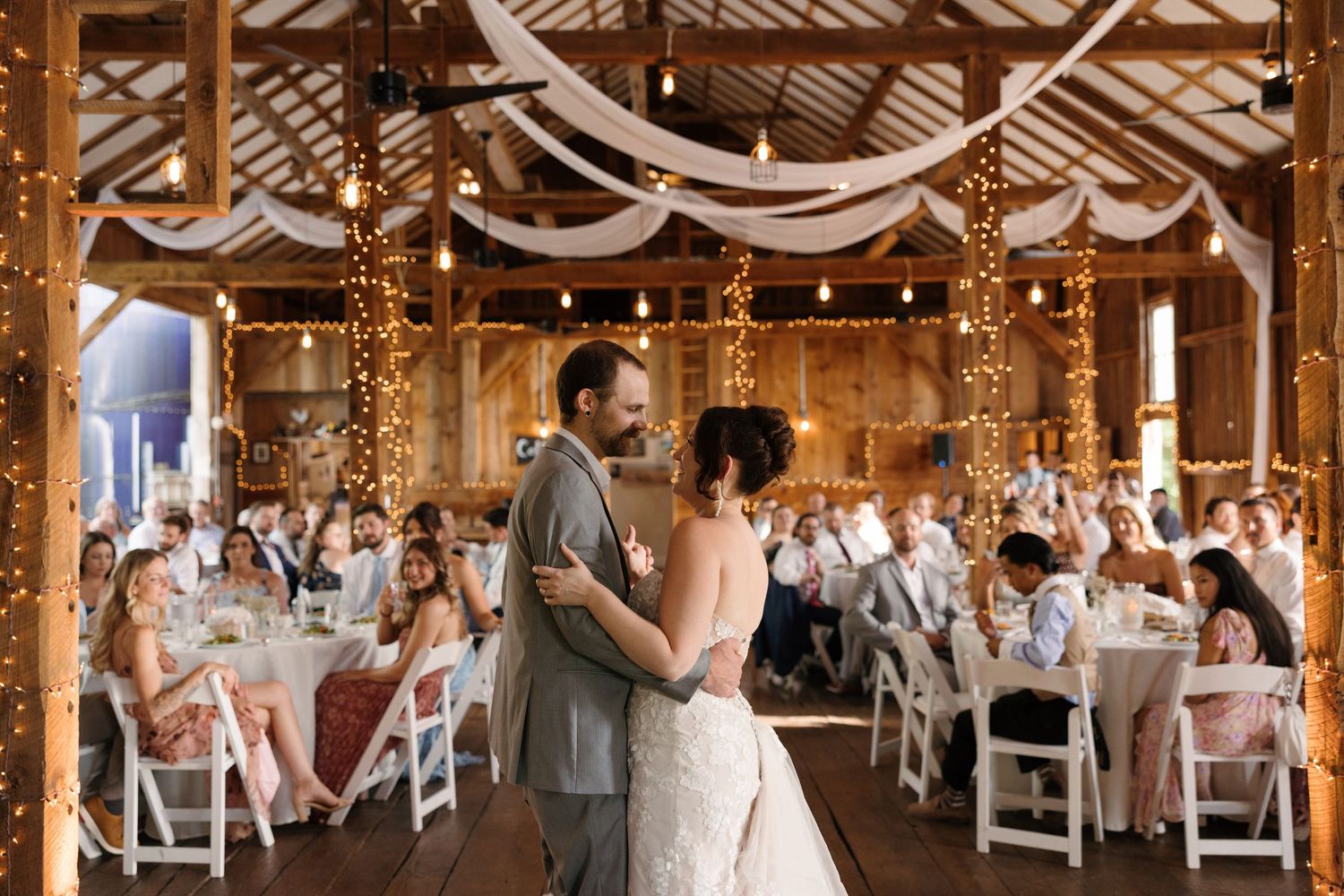 Bride and groom share first dance in a rustic barn wedding venue adorned with fairy lights and white draping.