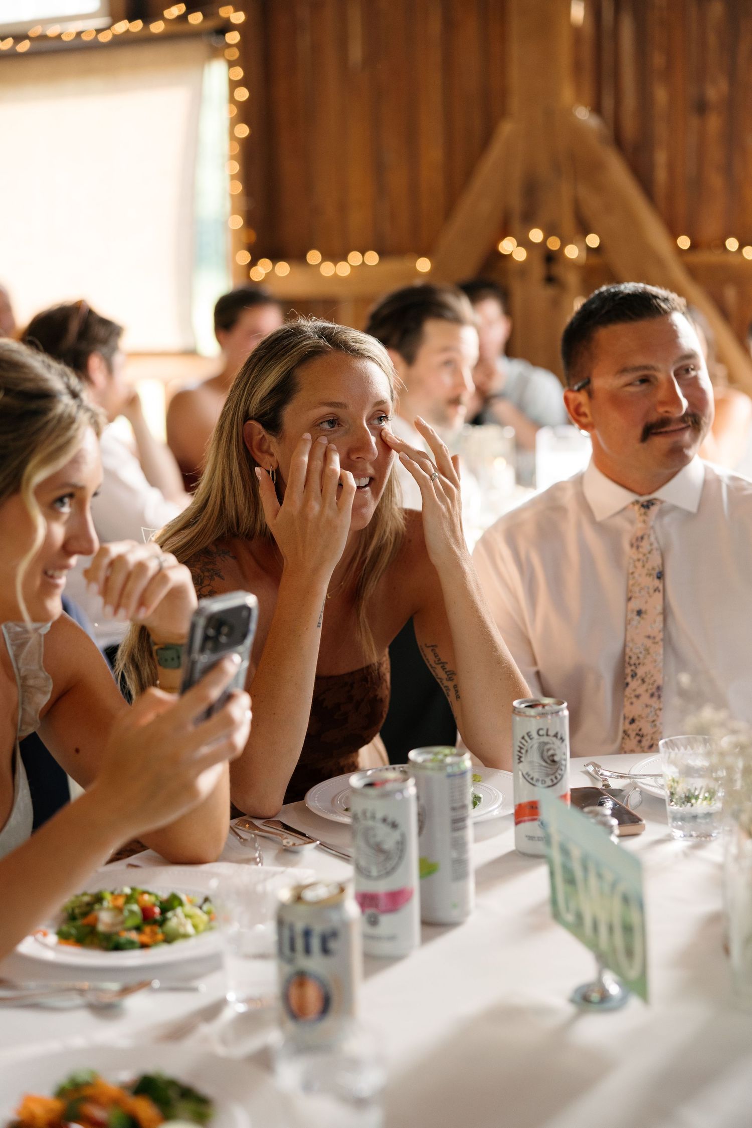 Guests laughing and crying at a wedding reception dinner in a rustic barn venue with string lights.