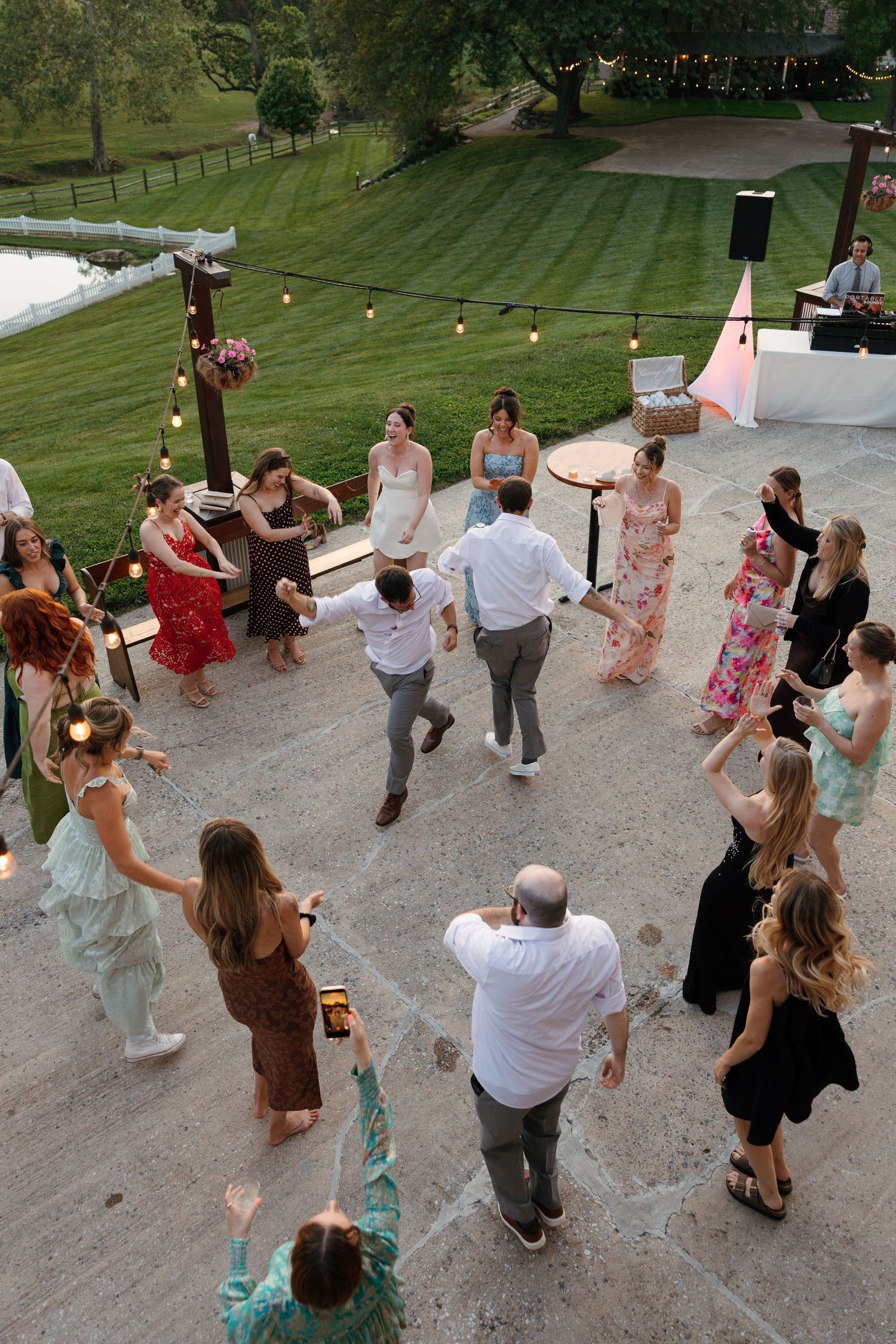 Guests dancing in a circle at an outdoor wedding reception on a patio surrounded by string lights and lush greenery.