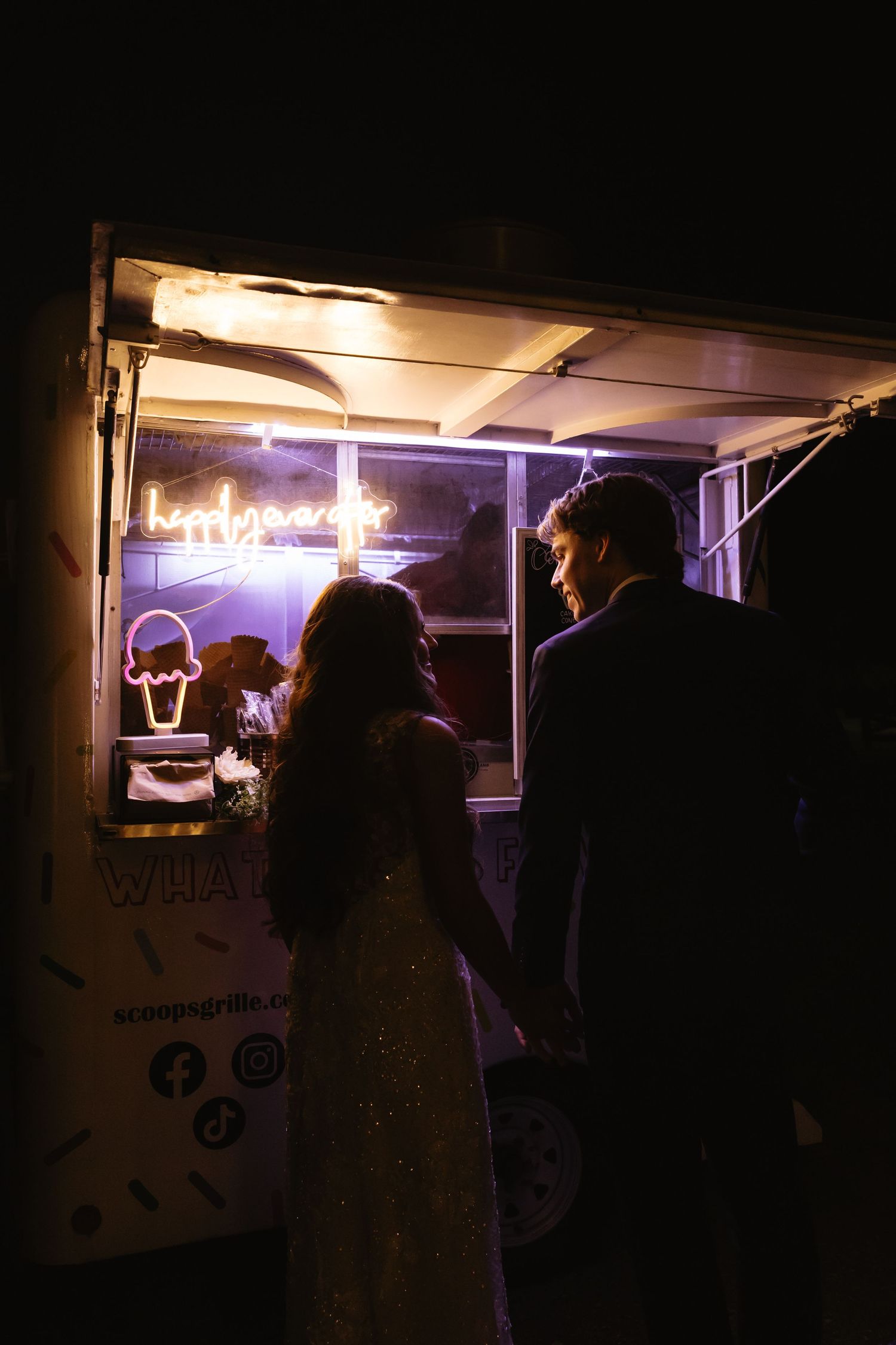 Silhouettes of customers line up at a food truck window glowing with purple neon light at night.