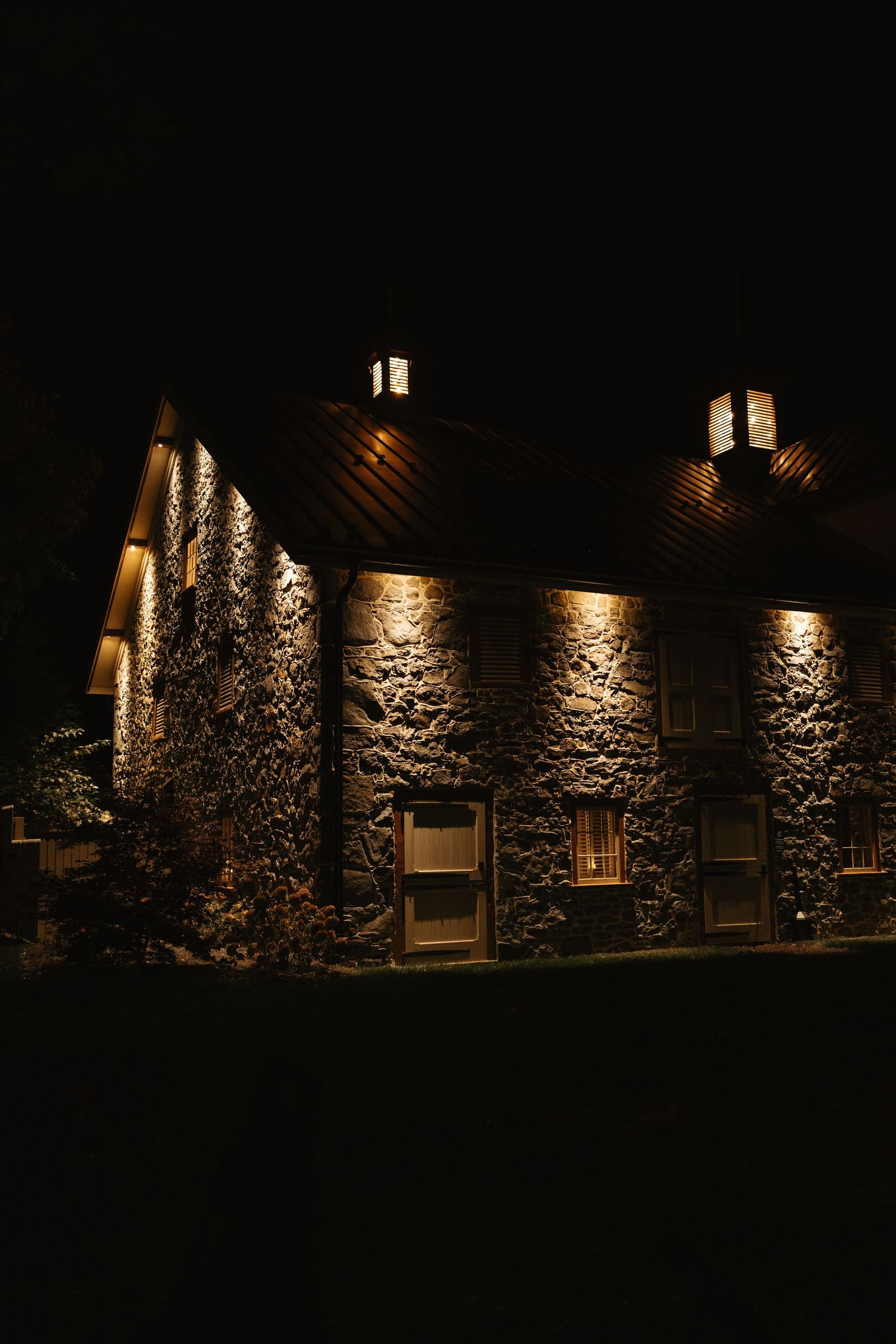 Stone wall building exterior illuminated at night with dramatic shadows and warm lighting across multiple windows.