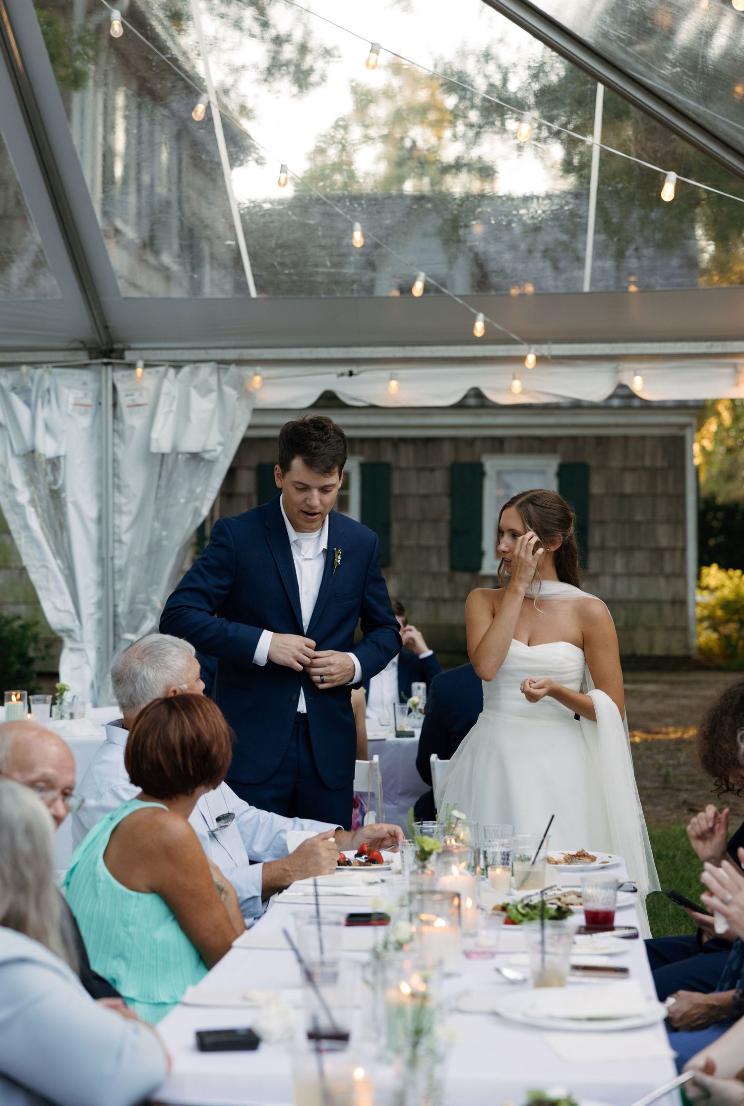 Wedding reception guests seated at elegant tables under a clear tent with hanging string lights and white decor.