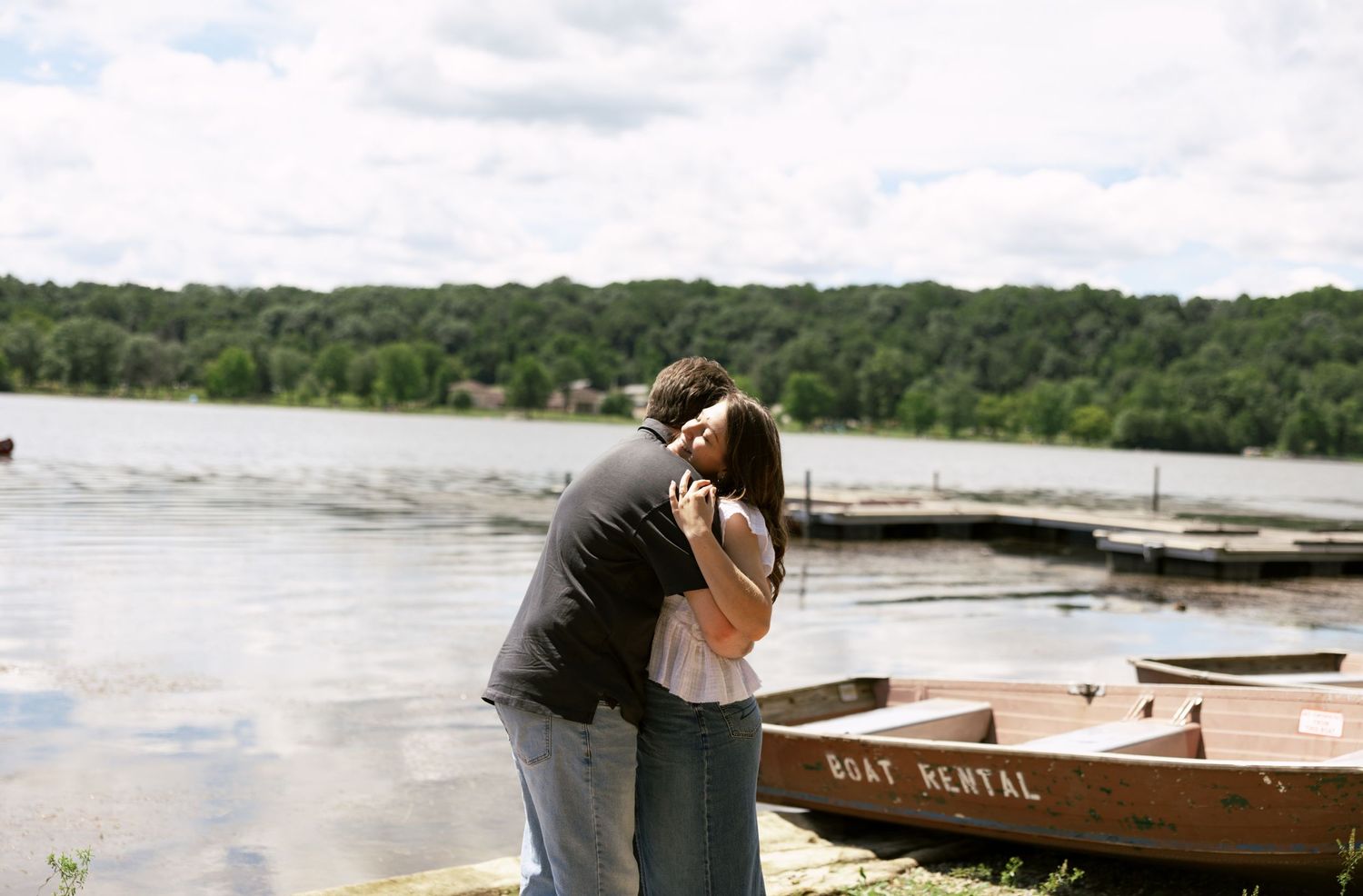 Couple sharing a romantic moment by a lake with wooden boats docked at a pier.