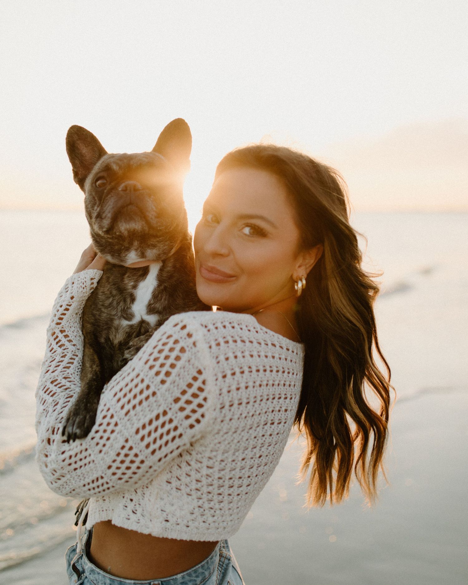 Woman in white crochet sweater holding French Bulldog on beach at sunset.