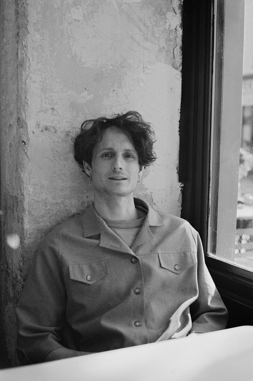 Black and white photograph of someone in a button-up jacket sitting by a window against a textured wall.