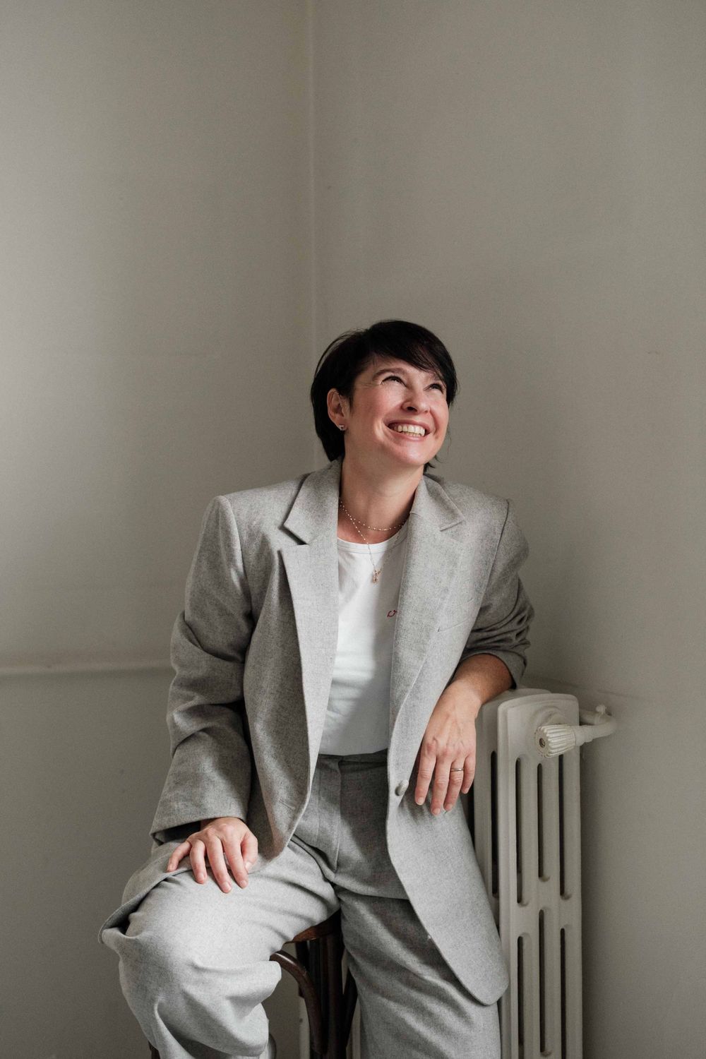 Professional portrait of someone in a grey suit leaning against a radiator with a bright smile.