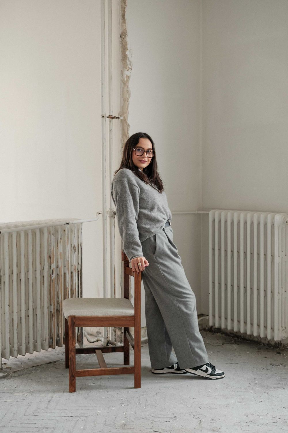 Person in grey loungewear standing beside a wooden chair against a white wall with vintage radiators.