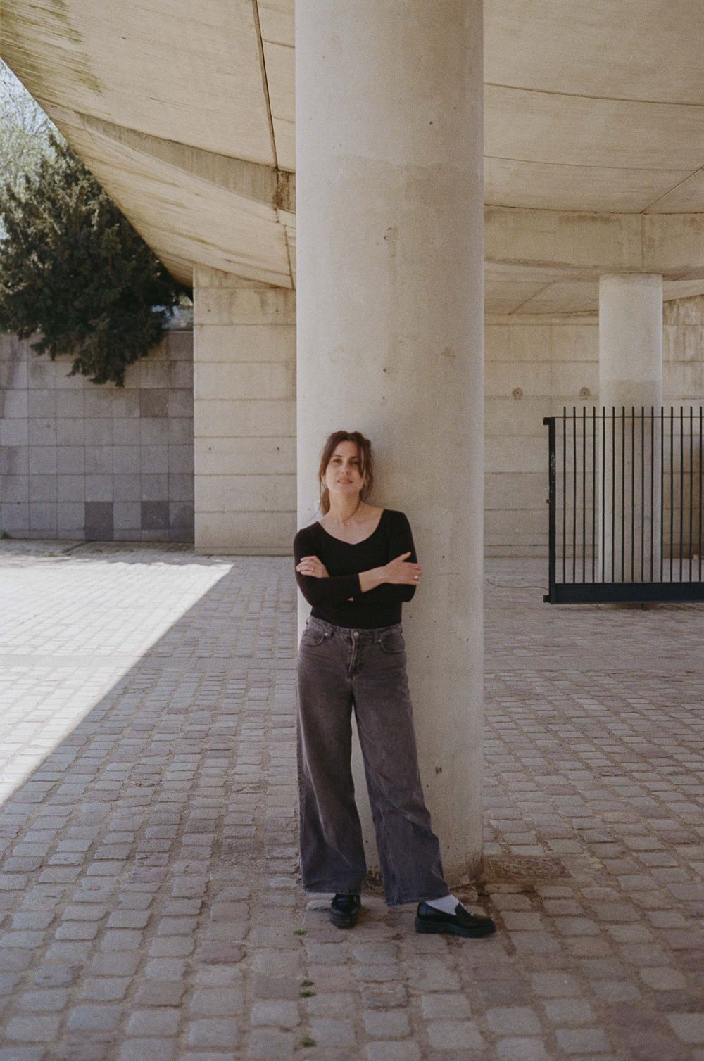 Person in casual black top and wide-leg jeans stands against white architectural column in urban courtyard setting.