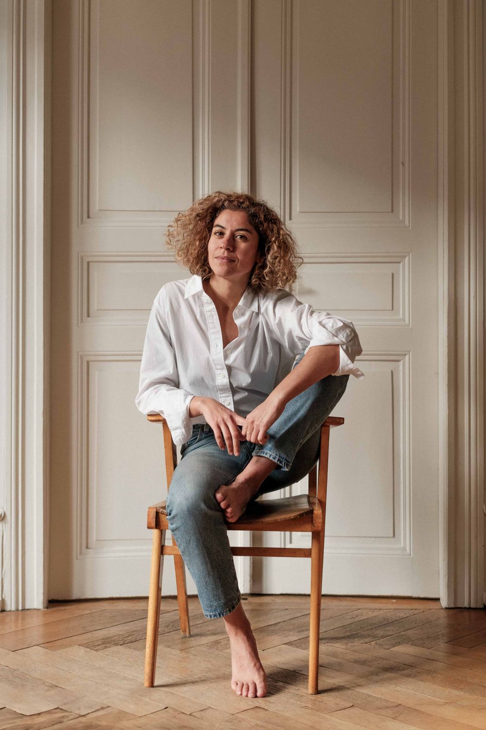 Casual portrait of someone in a white shirt and jeans sitting barefoot on a wooden chair against a paneled wall.