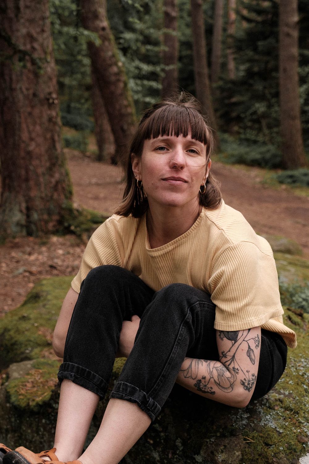 Casual portrait in forest setting with yellow t-shirt and visible tattoos.