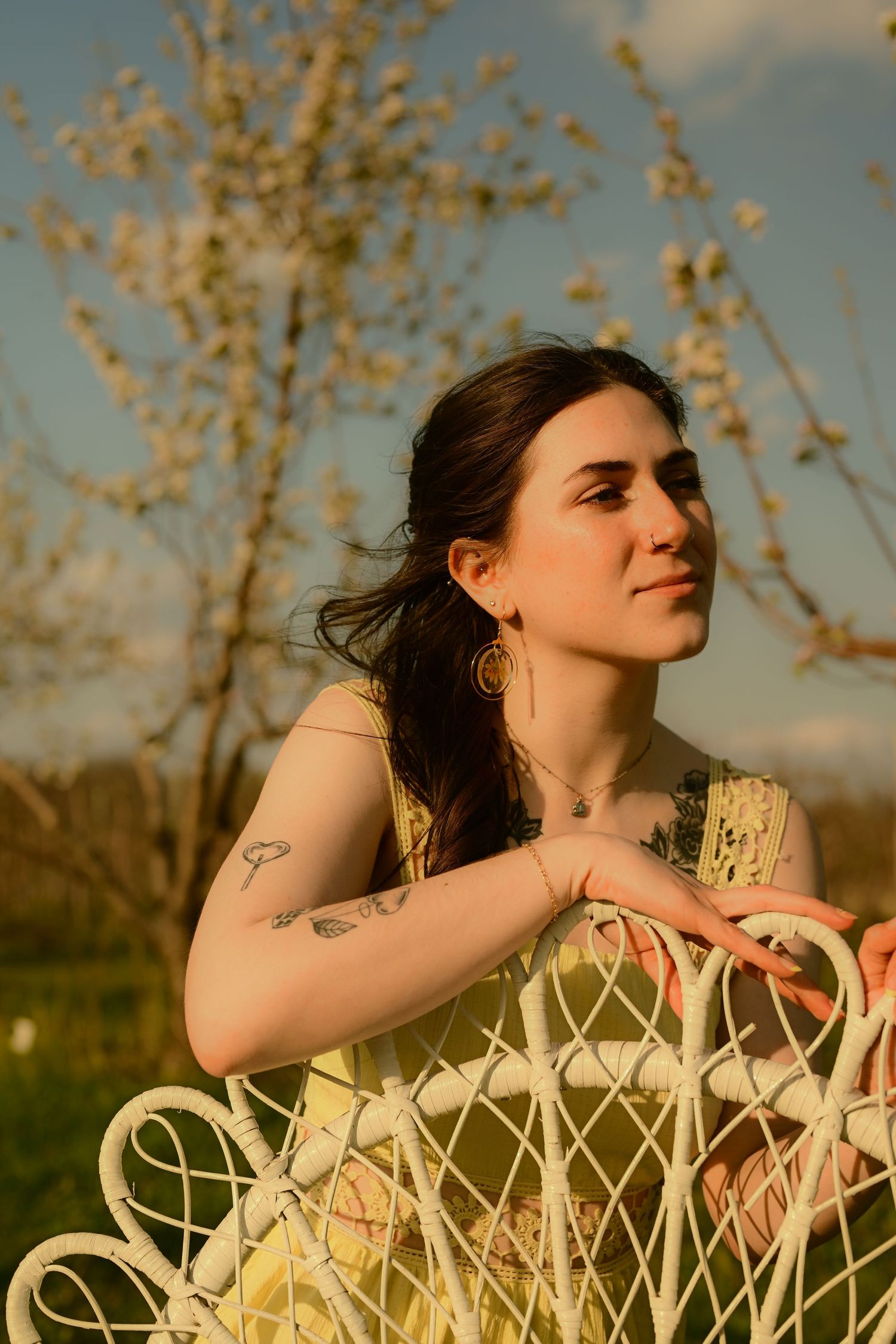 Young woman with tattoos in yellow dress leaning on white garden bench near blooming tree at golden hour.
