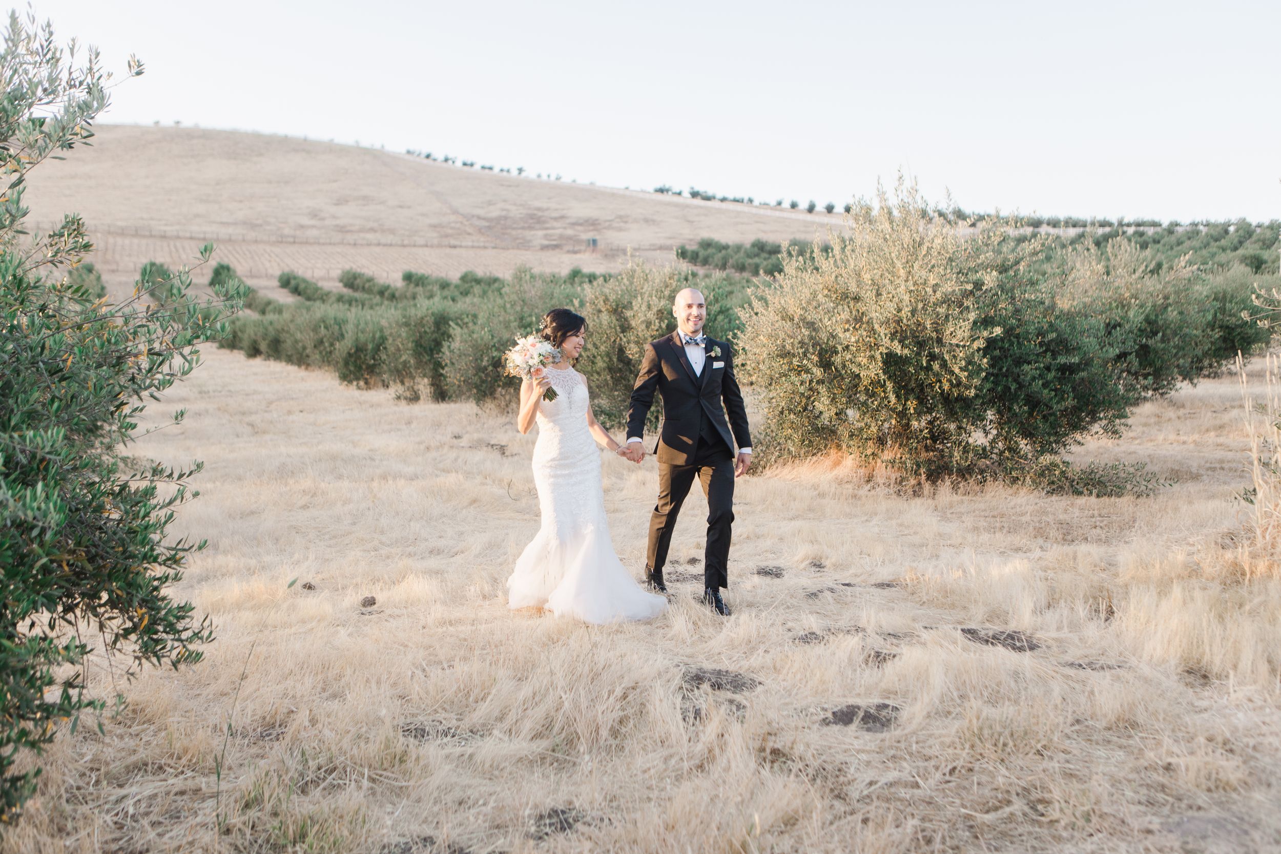 Bride and groom holding hands while walking through golden field at sunset.