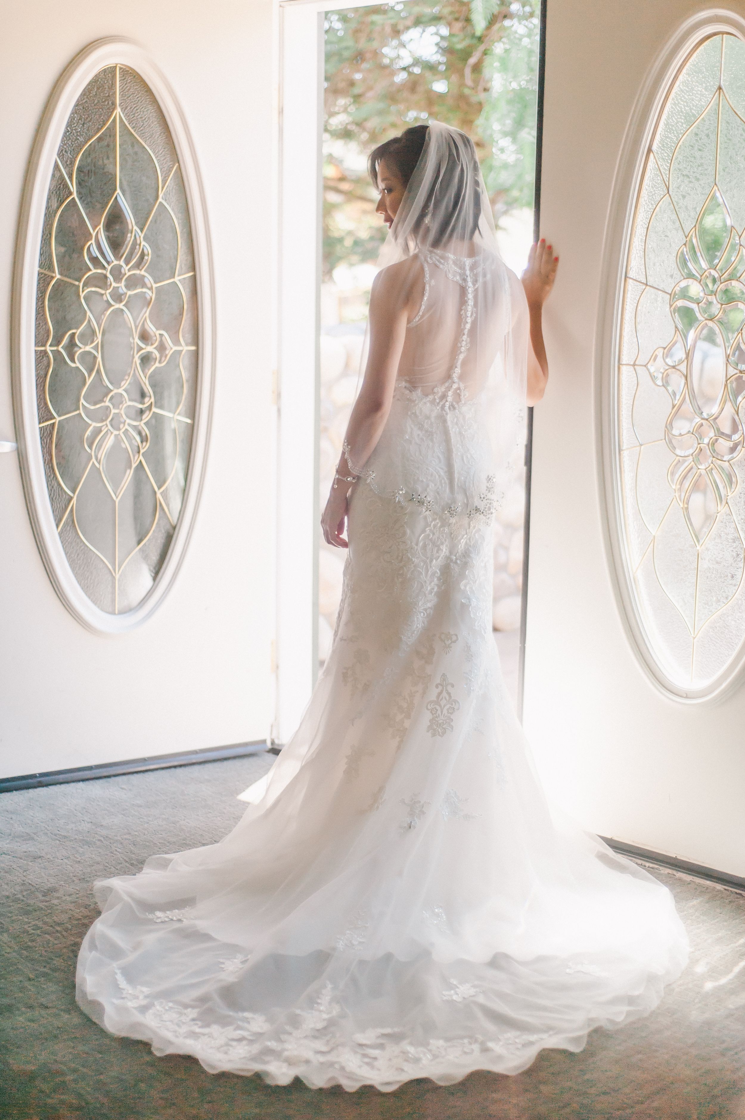 Bride in elegant lace wedding gown stands by doorway with decorative oval windows.