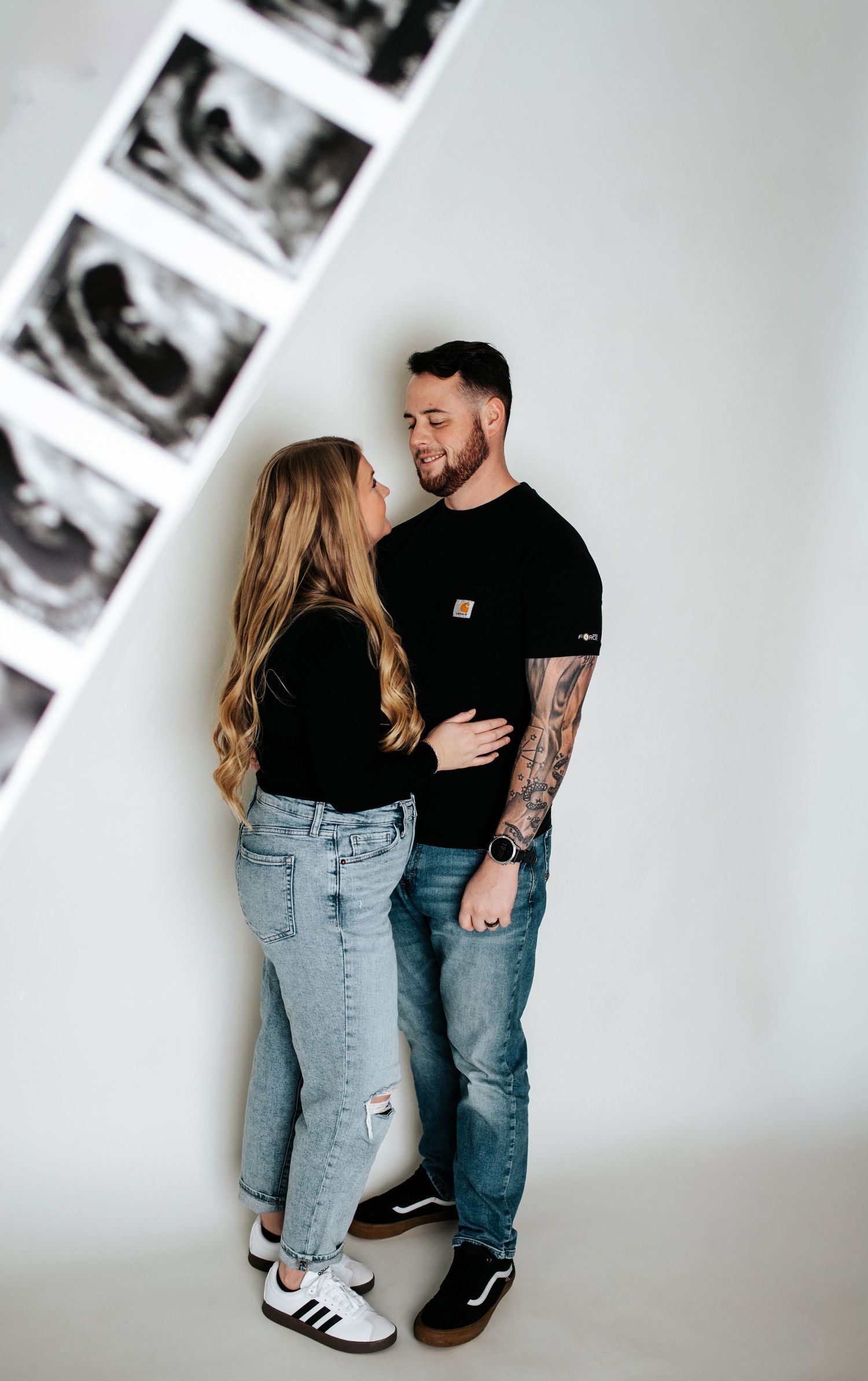 Couple in matching black outfits posing with ultrasound photos against white wall.