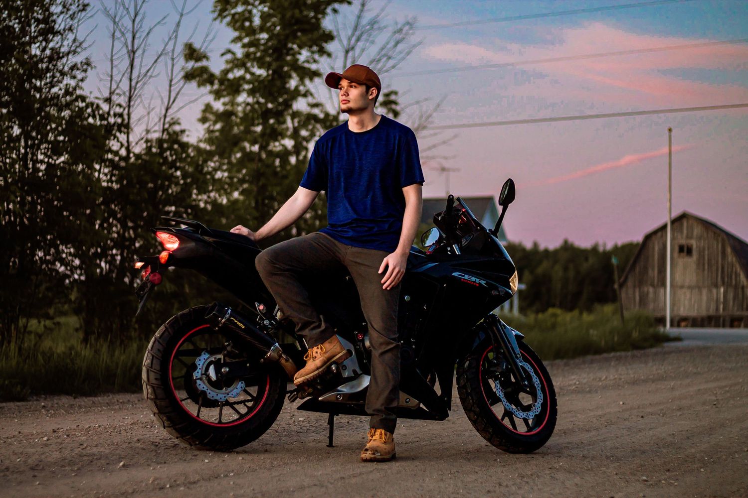 A motorcyclist in a blue shirt sits on a black sport bike at sunset with a barn in the background.