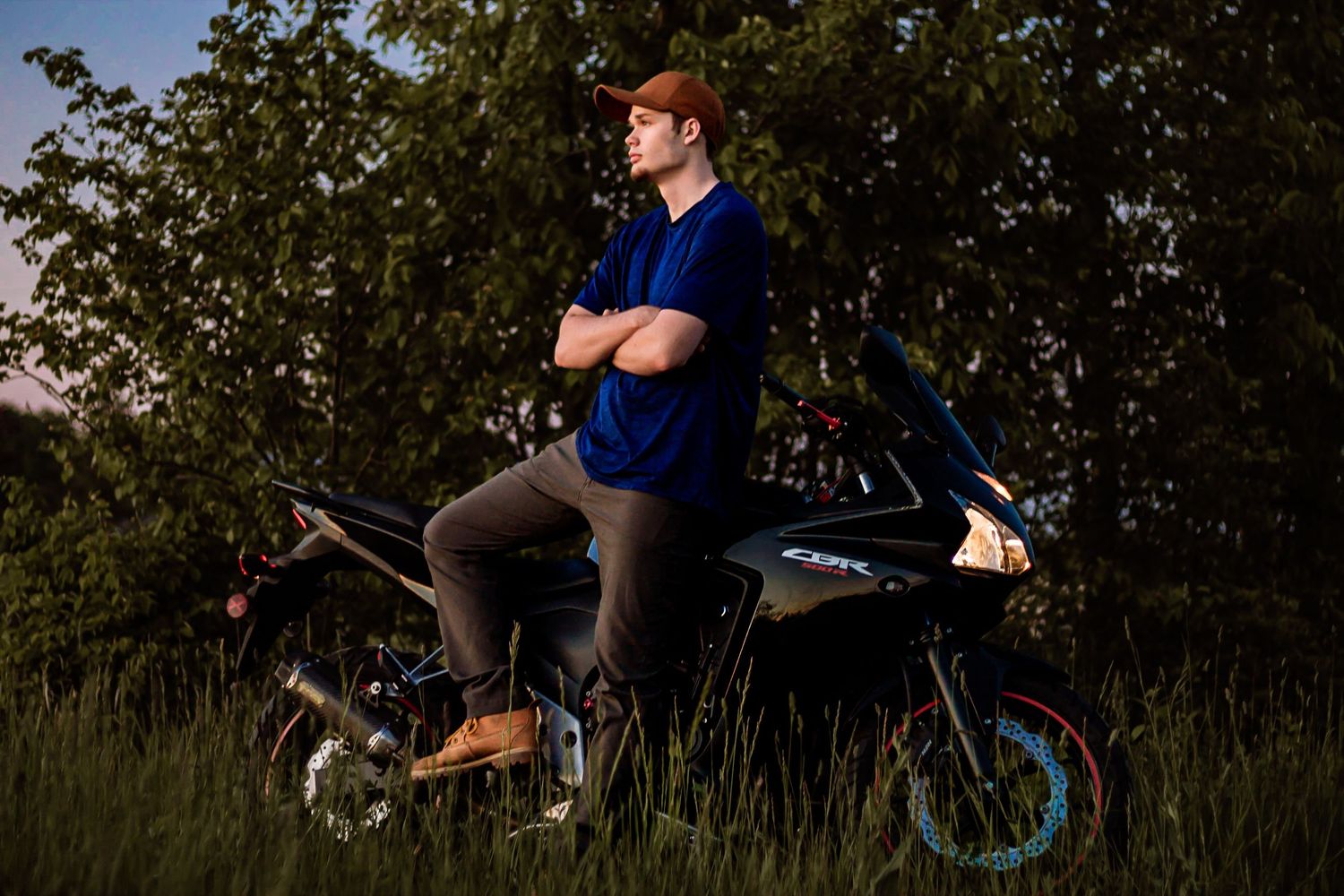Person stands confidently against sunset backdrop next to blue bicycle in grassy meadow with trees.