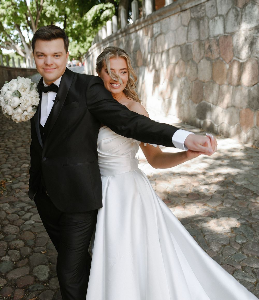 A couple in wedding attire dancing and twirling together on a stone path in dappled sunlight.