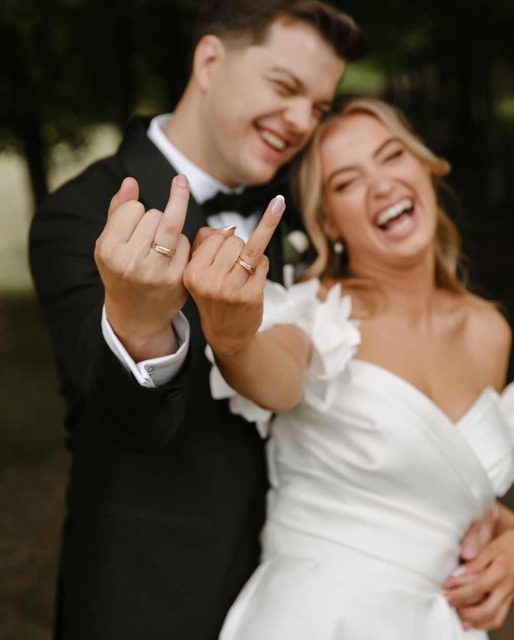 Joyful newlyweds laugh together while showing off their wedding rings in an outdoor portrait.