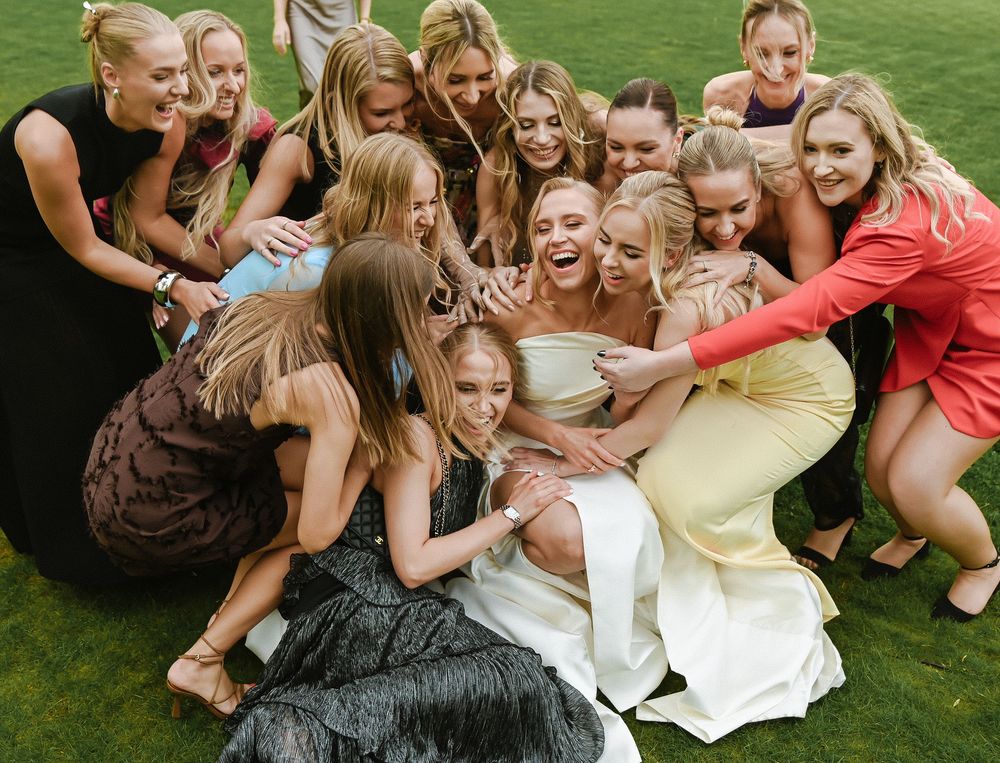Bridesmaids in colorful dresses huddle joyfully on grass for group photo.
