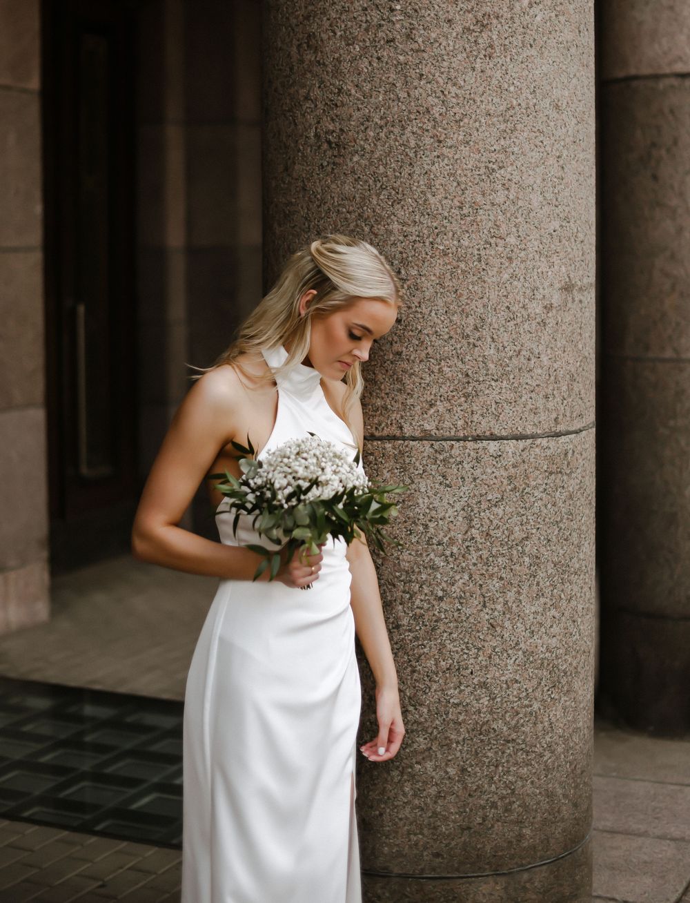 A bride in an elegant white halter dress holds a small bouquet while standing next to a stone pillar.