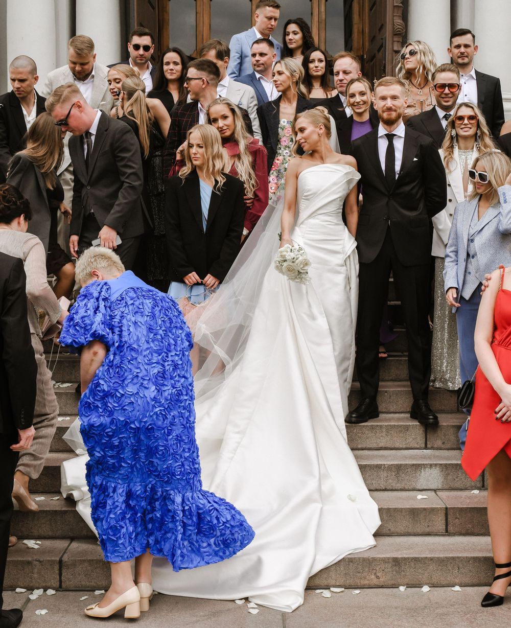Wedding party poses on historic church steps with bride in elegant white gown and guest in vibrant blue dress.