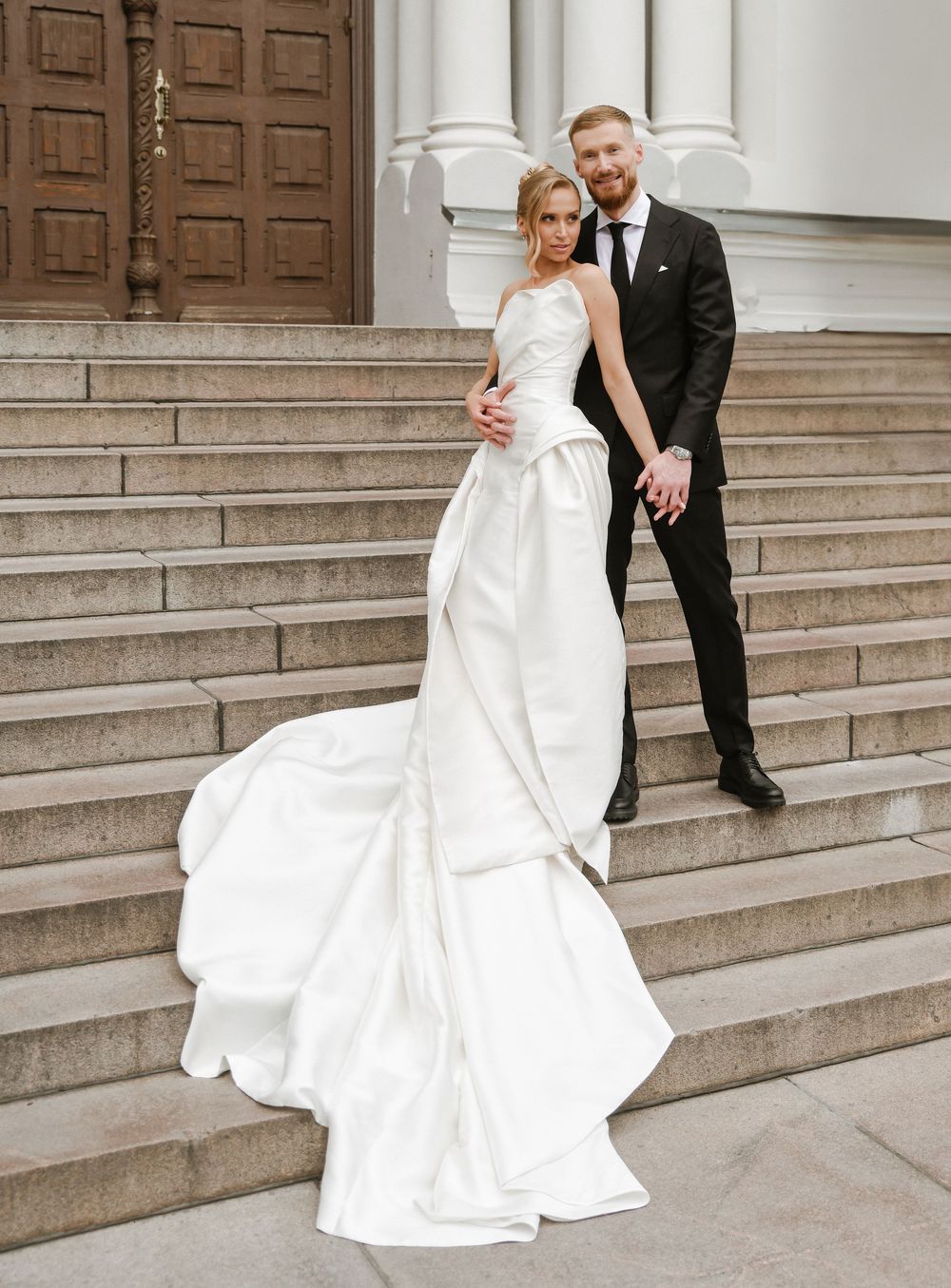 Elegant bride in flowing white gown poses with groom on grand stone church steps.
