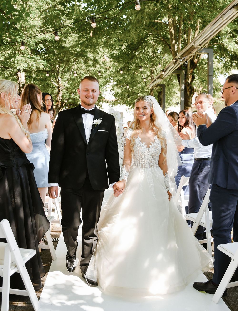 Wedding guests mingle and celebrate at an outdoor garden reception with white chairs under a tree canopy.