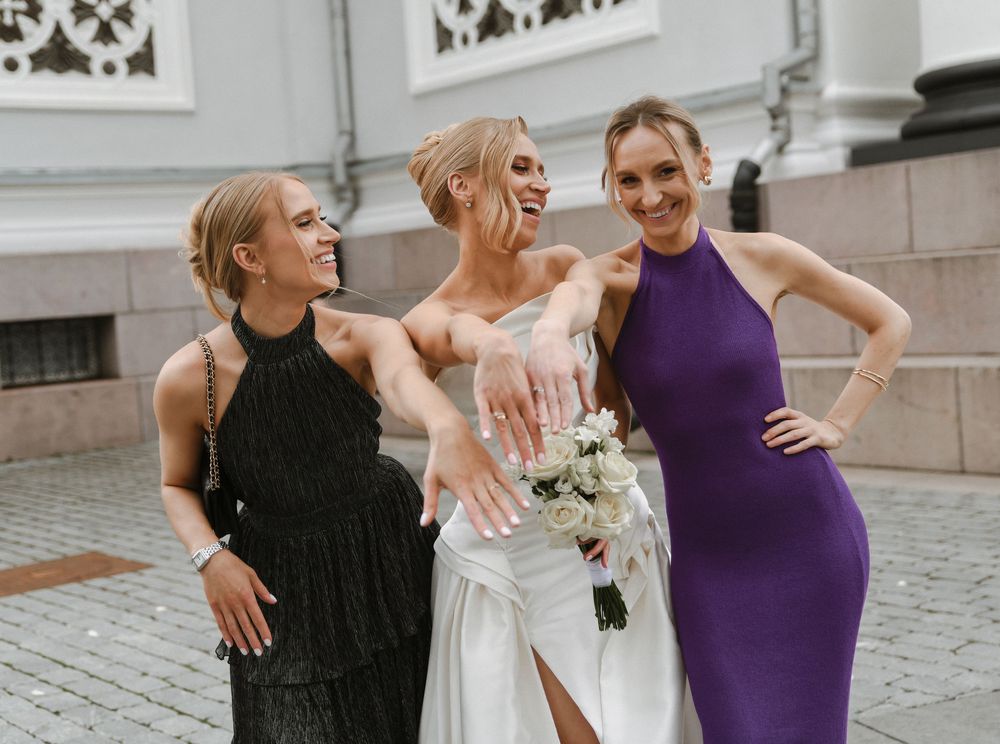Three people in formal attire laugh together outdoors, with one wearing a white wedding dress holding a bouquet.