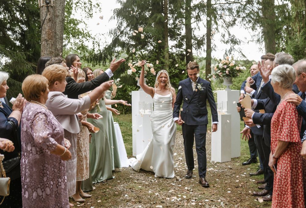 Wedding guests toss flower petals and confetti in celebration as they line both sides of an outdoor garden path.