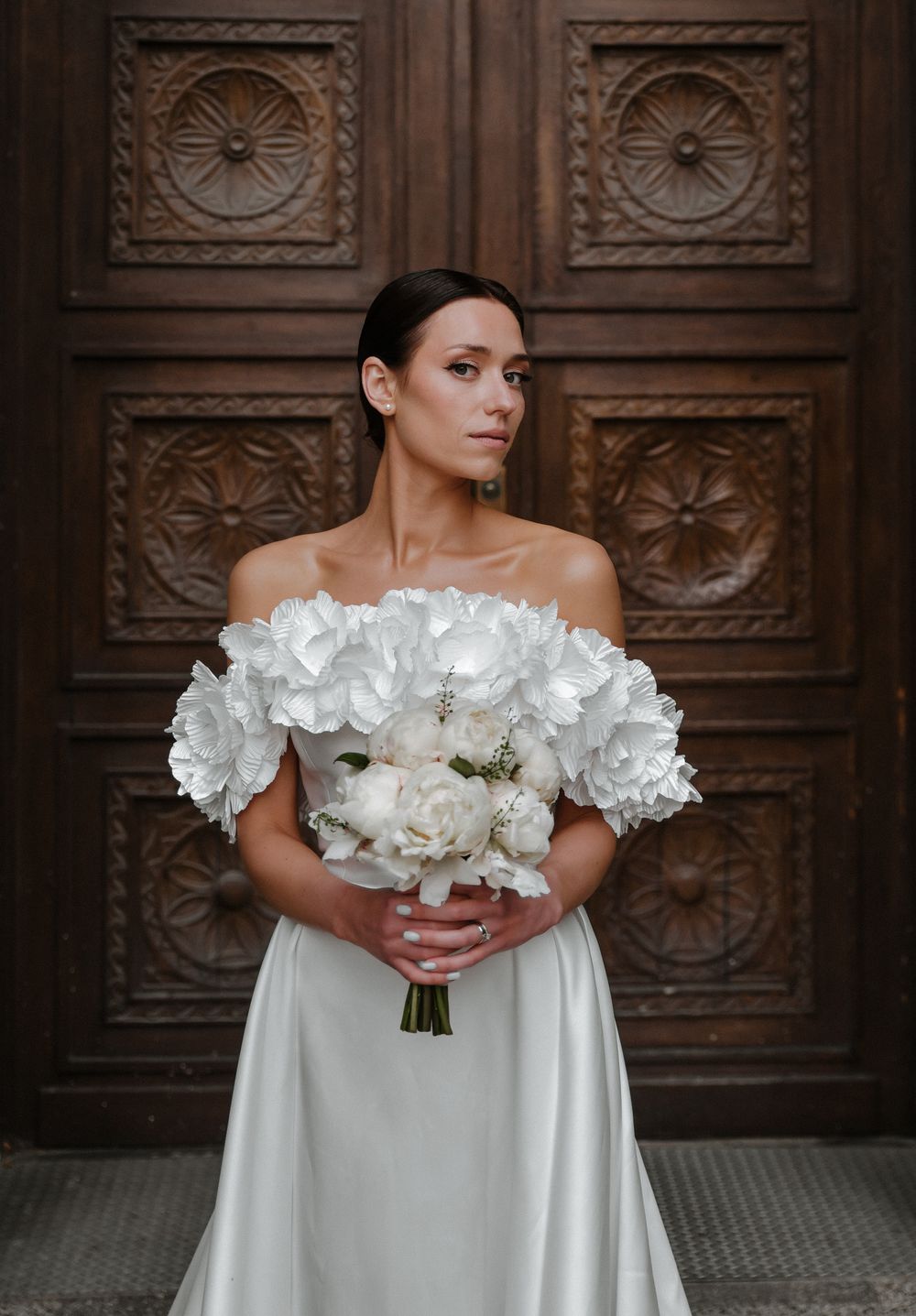 Bride in off-shoulder ruffled dress holds white bouquet against wooden door.