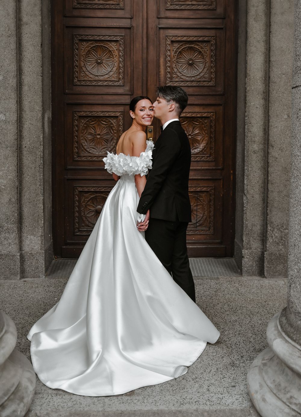 A romantic wedding portrait featuring an elegant off-shoulder white gown and black tuxedo against ornate wooden doors.