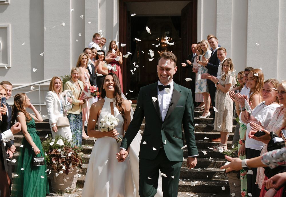 Newlyweds exit church while guests shower them with confetti on sunny day.
