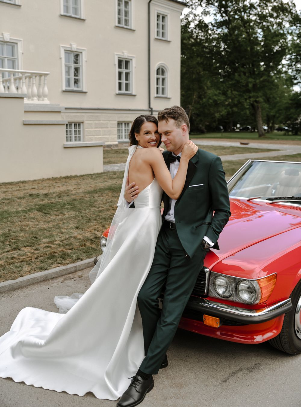 A wedding couple poses next to a classic red sports car outside a white building.