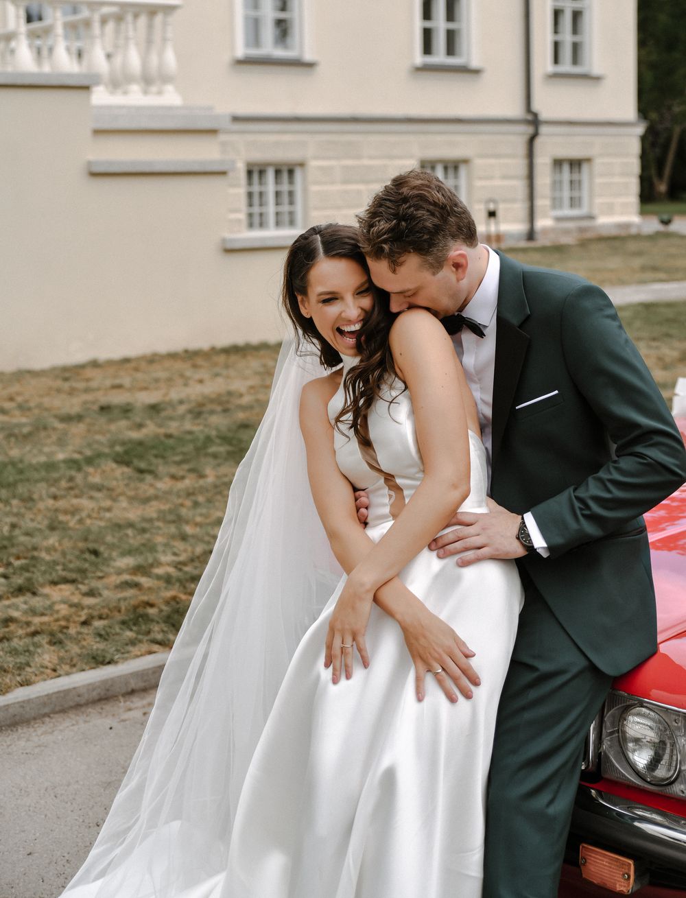 Newlyweds share a romantic moment beside a vintage red car.