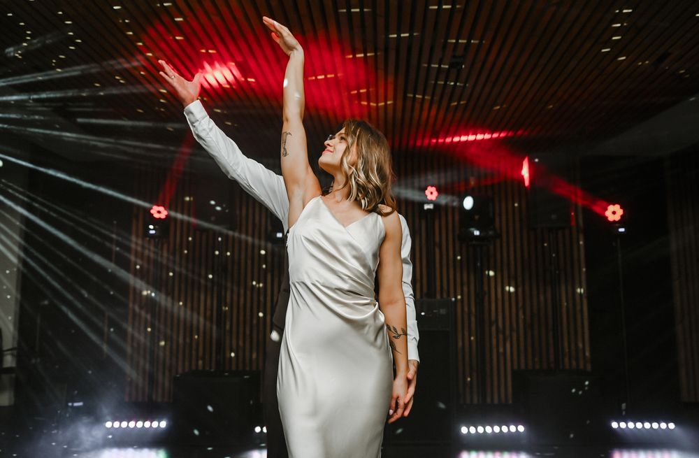 A concert performer in a white dress raises their arms under dramatic red stage lighting and illuminated string lights.