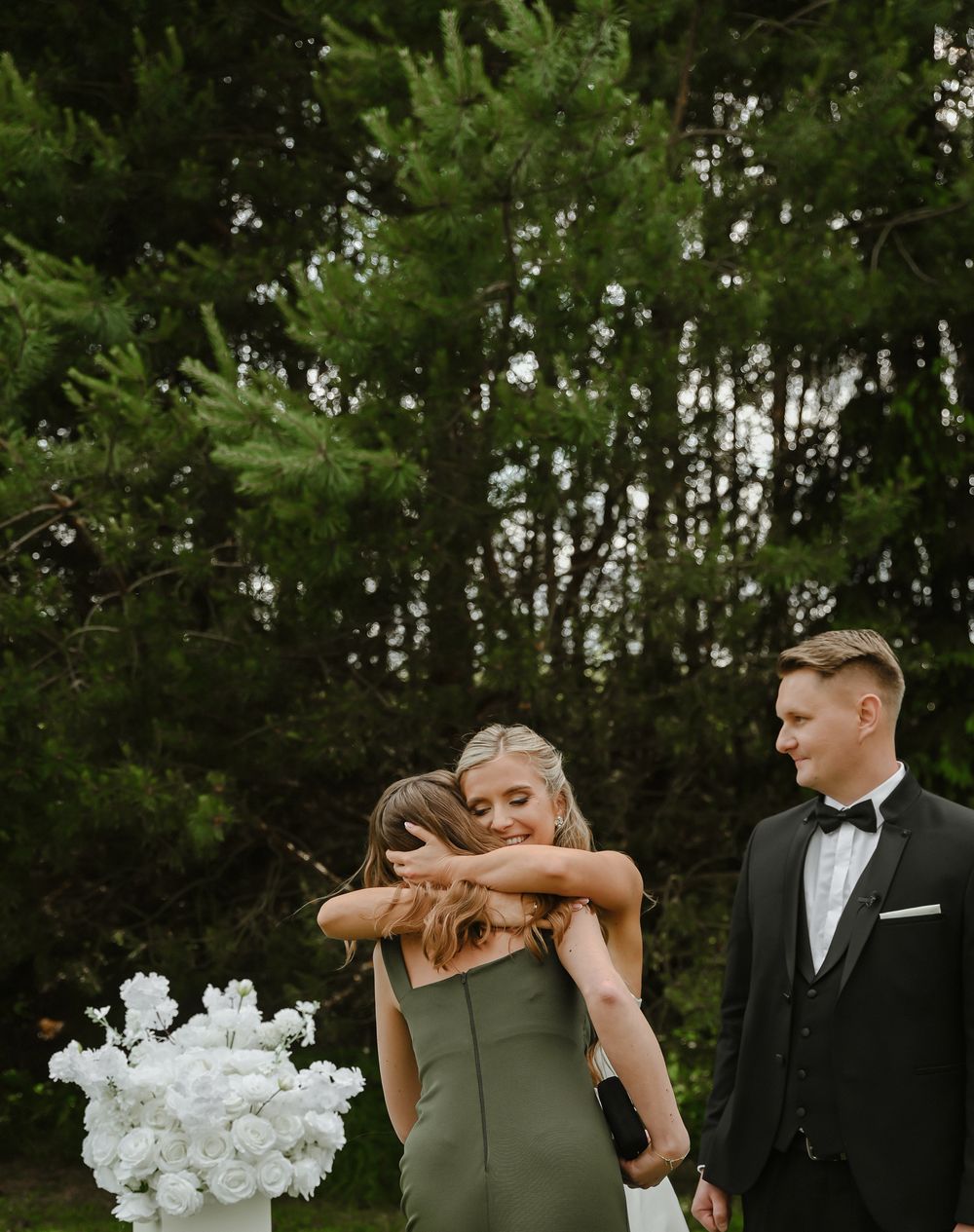 Wedding party in a forest setting with green bridesmaid dress and white floral bouquet against evergreen trees.