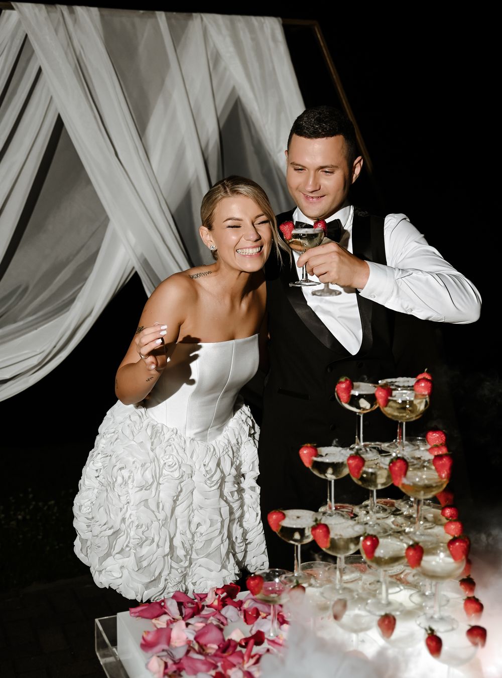 Wedding couple pouring champagne into a tower of glasses decorated with pink rose petals and strawberries.