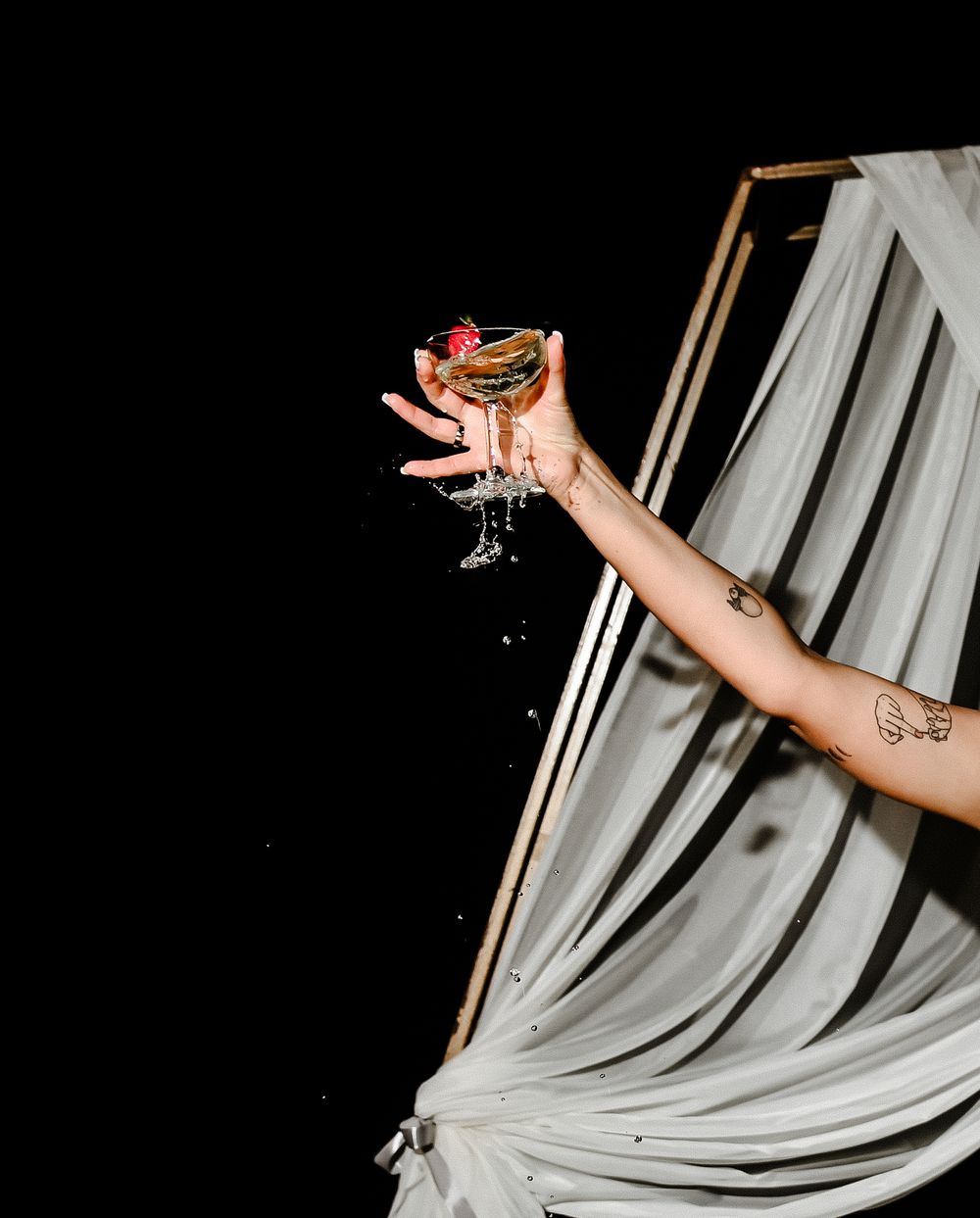 A hand holding up a martini glass with a pink cocktail against dark background and white draped fabric.